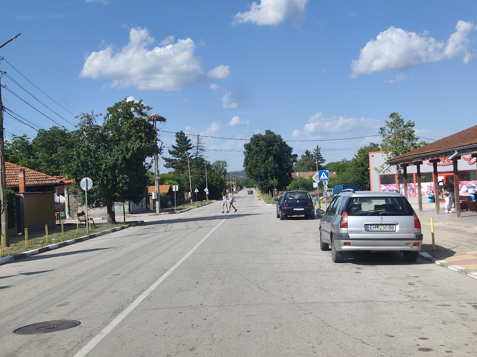 This is a street-level photograph taken in the village of Balgarene, Bulgaria, on a sunny day with a partly cloudy sky. The scene depicts a paved road running through a village with buildings and trees lining its sides. In the foreground, a silver Peugeot station wagon is parked on the right side of the road. Further down the road, two individuals are crossing from left to right at a pedestrian crossing. On the left side of the road, there are houses with red tile roofs and a street lamp. On the right side, there is a building with a covered outdoor seating area, possibly a cafe or shop. In the distance, more buildings and trees are visible, with a stork's nest on a pole in the upper left portion of the image. The road has painted white lines indicating lanes and a white line marking the edge of the road.