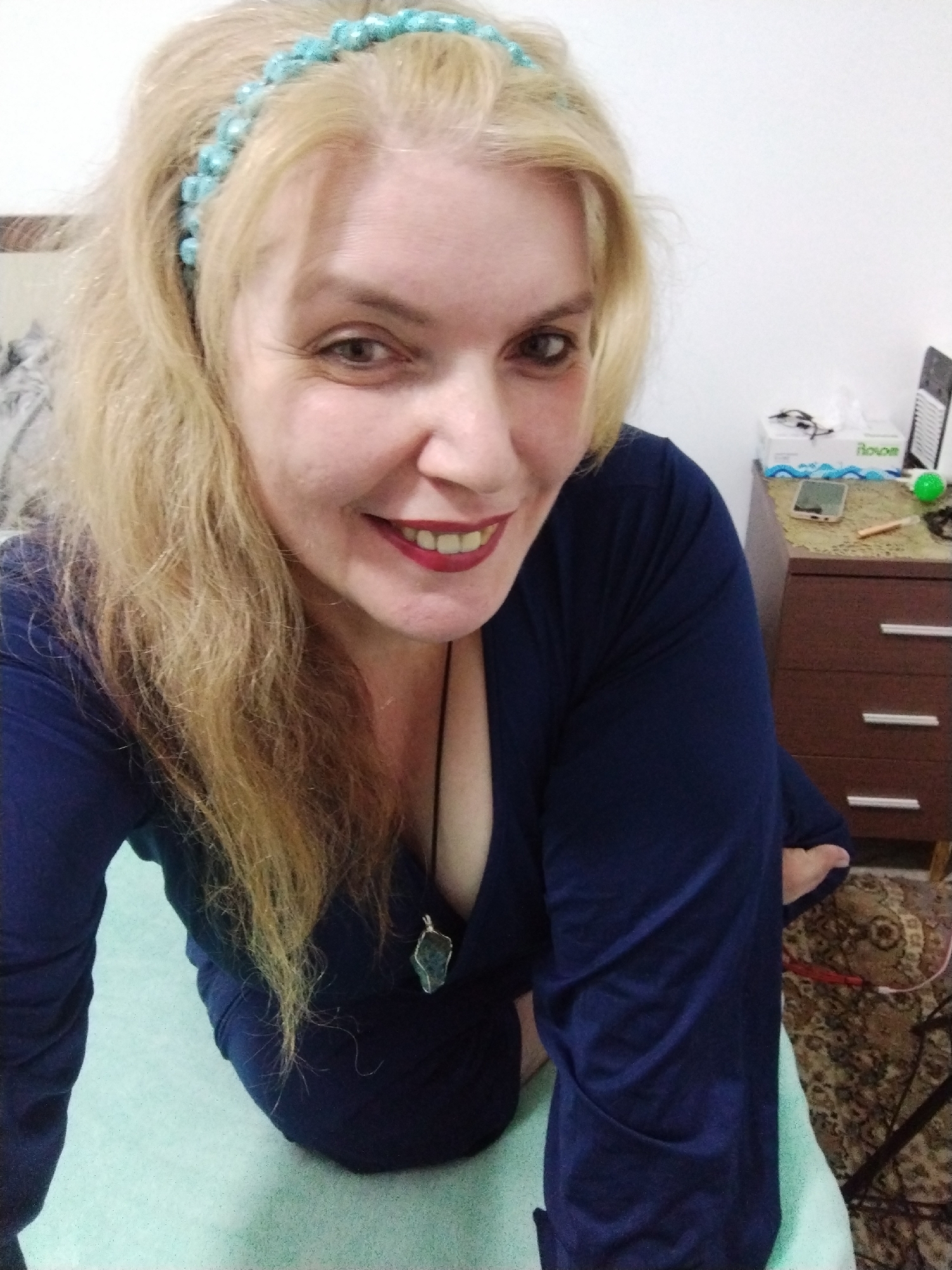 A woman with blonde hair and a turquoise beaded headband is kneeling on a light green surface. She is wearing a dark blue, long-sleeved, V-neck dress and a necklace with a light-colored pendant. She is smiling at the camera with red lipstick. Behind her is a brown wooden dresser with three drawers, a box of tissues, a cell phone, and other objects on top. The background also shows a patterned rug. The location is possibly inside a home or apartment building, but the exact address or broader city area (Al Mas'oudi, United Arab Emirates) cannot be confirmed from the image alone.