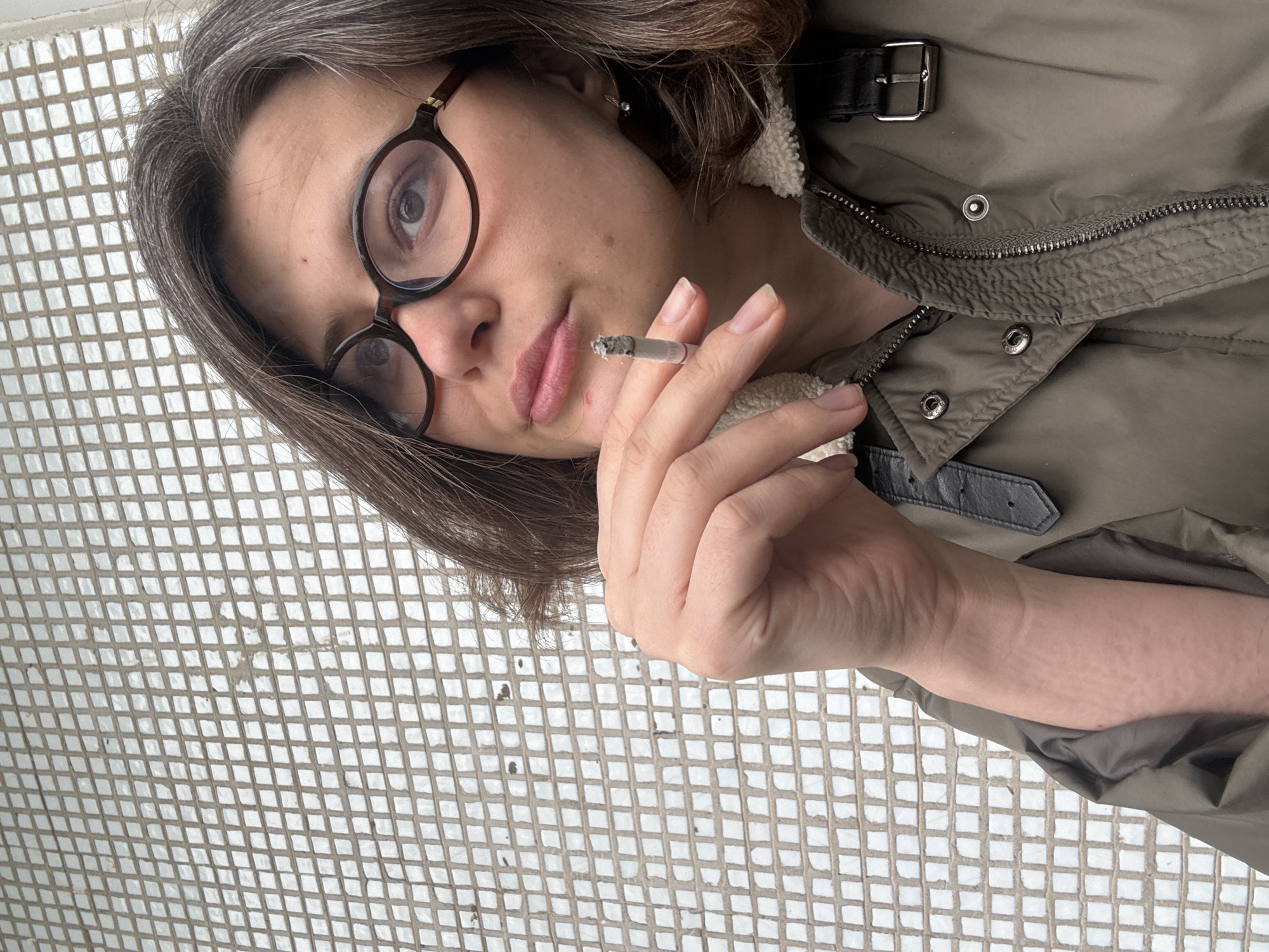 A woman with grey-streaked brown hair and round-frame glasses holds a lit cigarette near her mouth. She is wearing a green jacket with a sherpa-lined collar and black leather details, including a buckle closure on the collar. The background appears to be a wall covered in small, square, white tiles. The location is not discernible from the image and cannot be specifically identified as being in the city of Chaikovsky, Russia.