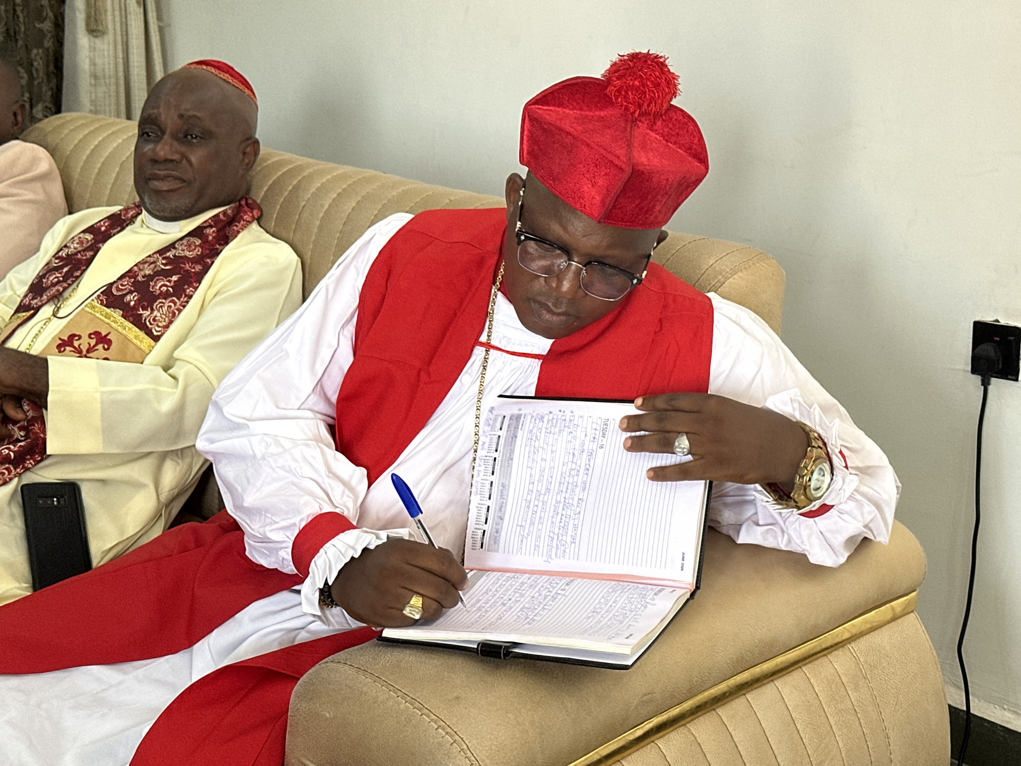 A man wearing a red clerical hat, a white shirt with red trim, and a red vest is seated on a tan couch. He is wearing glasses and a gold chain. He is writing in a notebook with a blue pen. On his left hand, he wears a gold ring, and on his right wrist, he wears a gold watch. To his left, another man in similar attire with a red and gold patterned scarf is seated. A black smartphone is visible next to him. The background is a plain white wall. The scene appears to be indoors in Narayi, Nigeria.