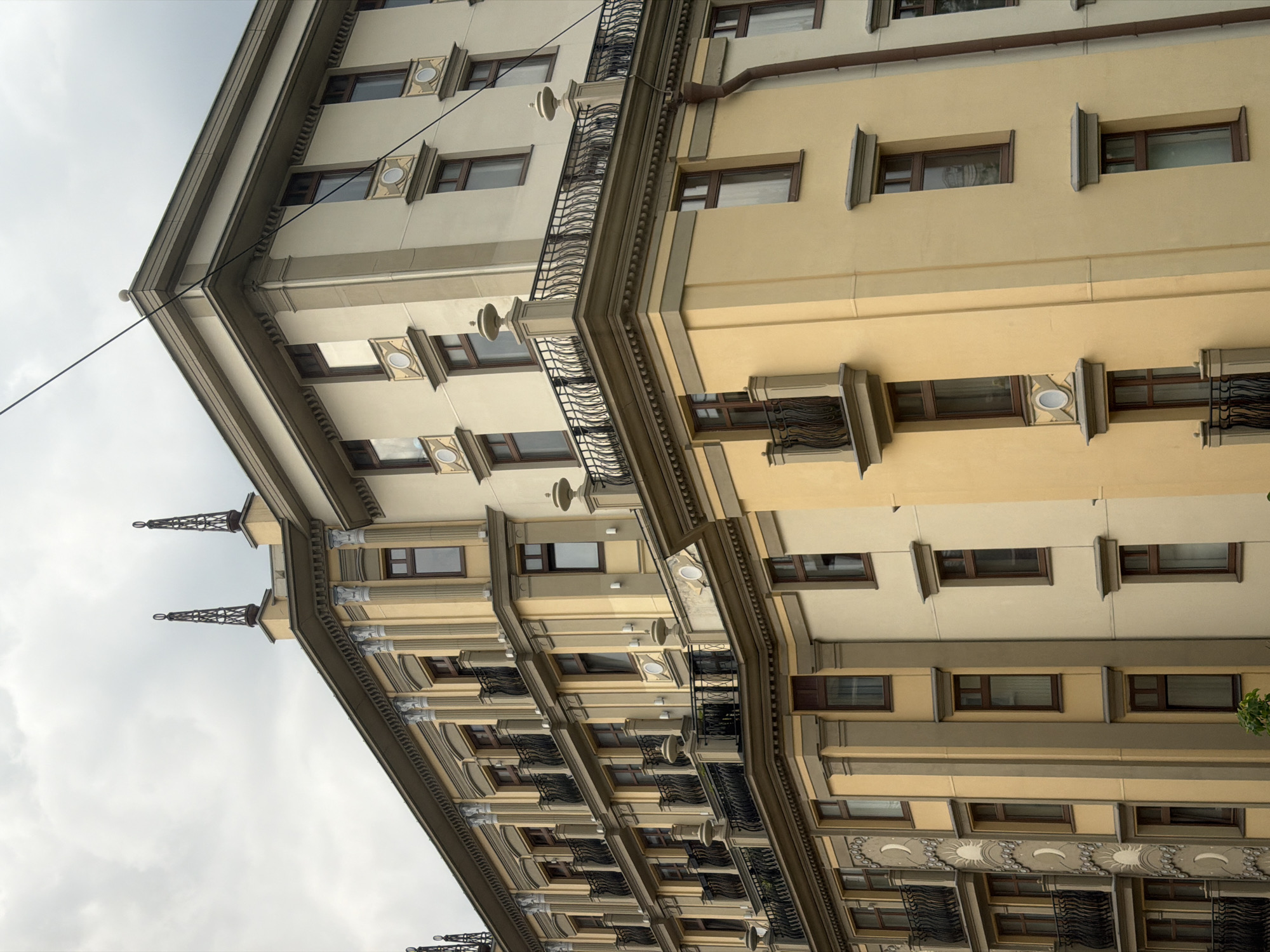 The image shows a tall, multi-story building in Moscow, Russia, likely an apartment complex, viewed from a low angle. The building has ornate architectural details, including protruding window frames, decorative balconies with black metal railings, and a two-toned color scheme of beige and yellow. Two spires extend from the roof of the building in a grey sky. A powerline crosses the image. The perspective is rotated counterclockwise.