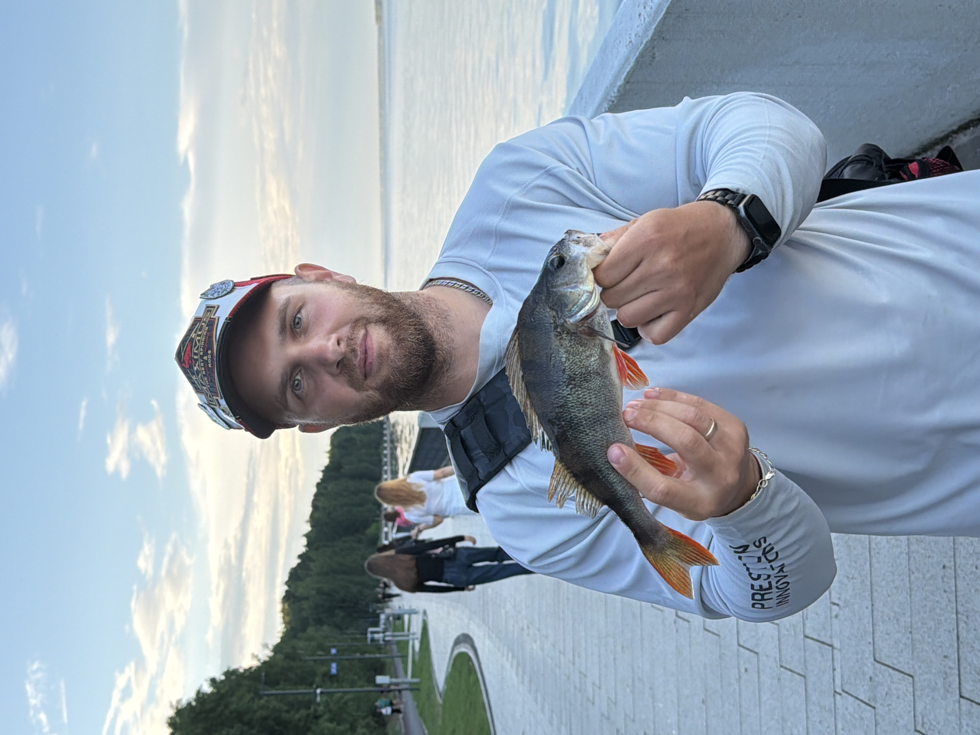 A man stands on a waterfront promenade in Chaikovsky, Russia, holding a freshly caught perch. He is wearing a light gray long-sleeved shirt with "PRESTON INNOVATIONS" printed on the left sleeve, a black strap across his shoulders, and a baseball cap. The man has a beard and is looking at the camera. Behind him, a paved walkway curves along the shore, with trees and a body of water to his right. Several people are walking on the walkway in the background. The sky is partly cloudy.