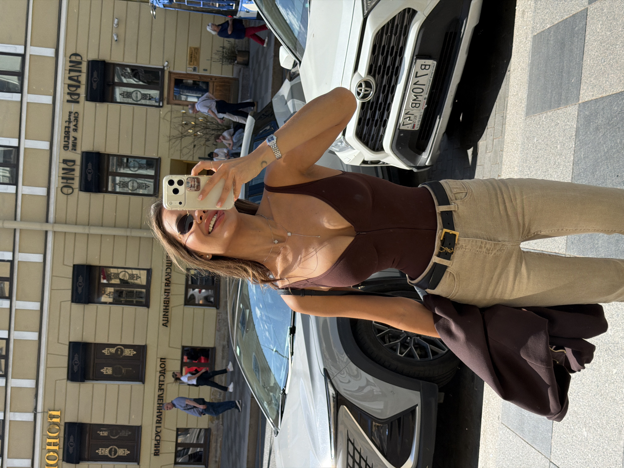 The image shows a woman taking a selfie on a sunny day in Saint Petersburg, Russia. She is wearing a brown bodysuit, beige pants, and a black belt with a gold YSL buckle. She is also holding a dark brown jacket over her left arm and wearing sunglasses and a necklace. The background features a yellow building with ornate windows and signage that appears to be in Russian. Two cars are parked on the street, one silver and one white. Several people are walking in the background, some near the building and others on the sidewalk. The scene is set on a paved street with visible stone tiling.