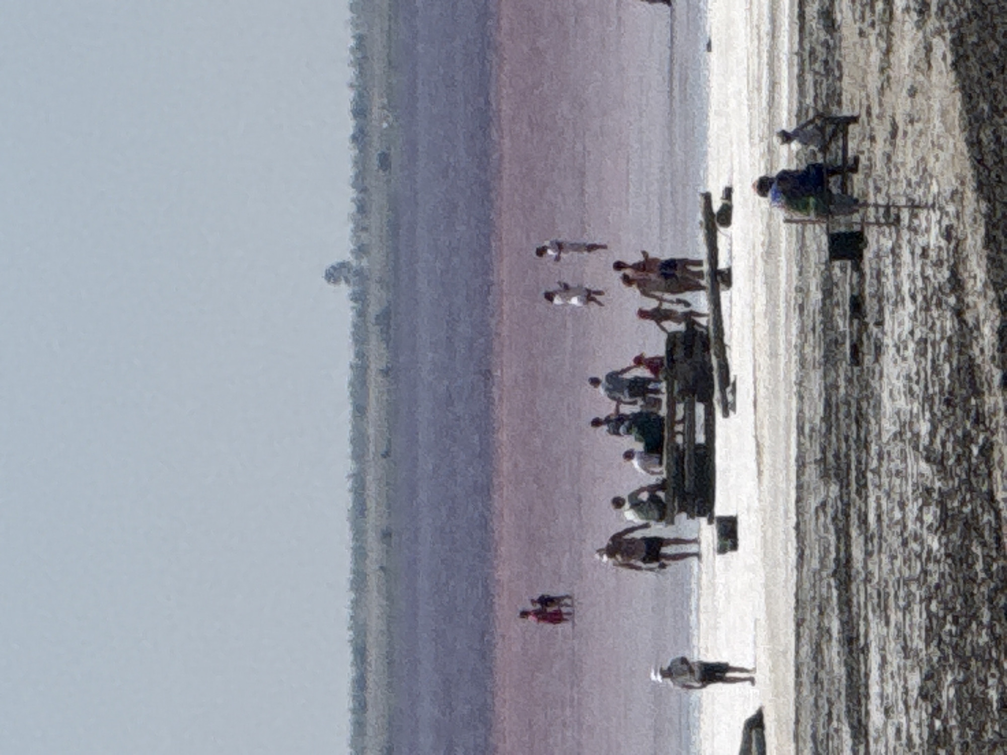 This image shows a view of a salt lake in Bursol, Russia, on a sunny day. The water of the lake has a distinct pinkish hue. In the foreground, there is a sandy or muddy shore littered with debris. Several people are visible in and around the water. Some are wading, others are standing or sitting on the shore. One group is gathered around a wooden table, appearing to be relaxing or having a picnic. Another person is seen standing on the shore wearing a hat and shorts. In the background, a thin strip of land with some vegetation separates the pink lake from a hazy blue sky. The overall impression is of a recreational area near a salt lake.
