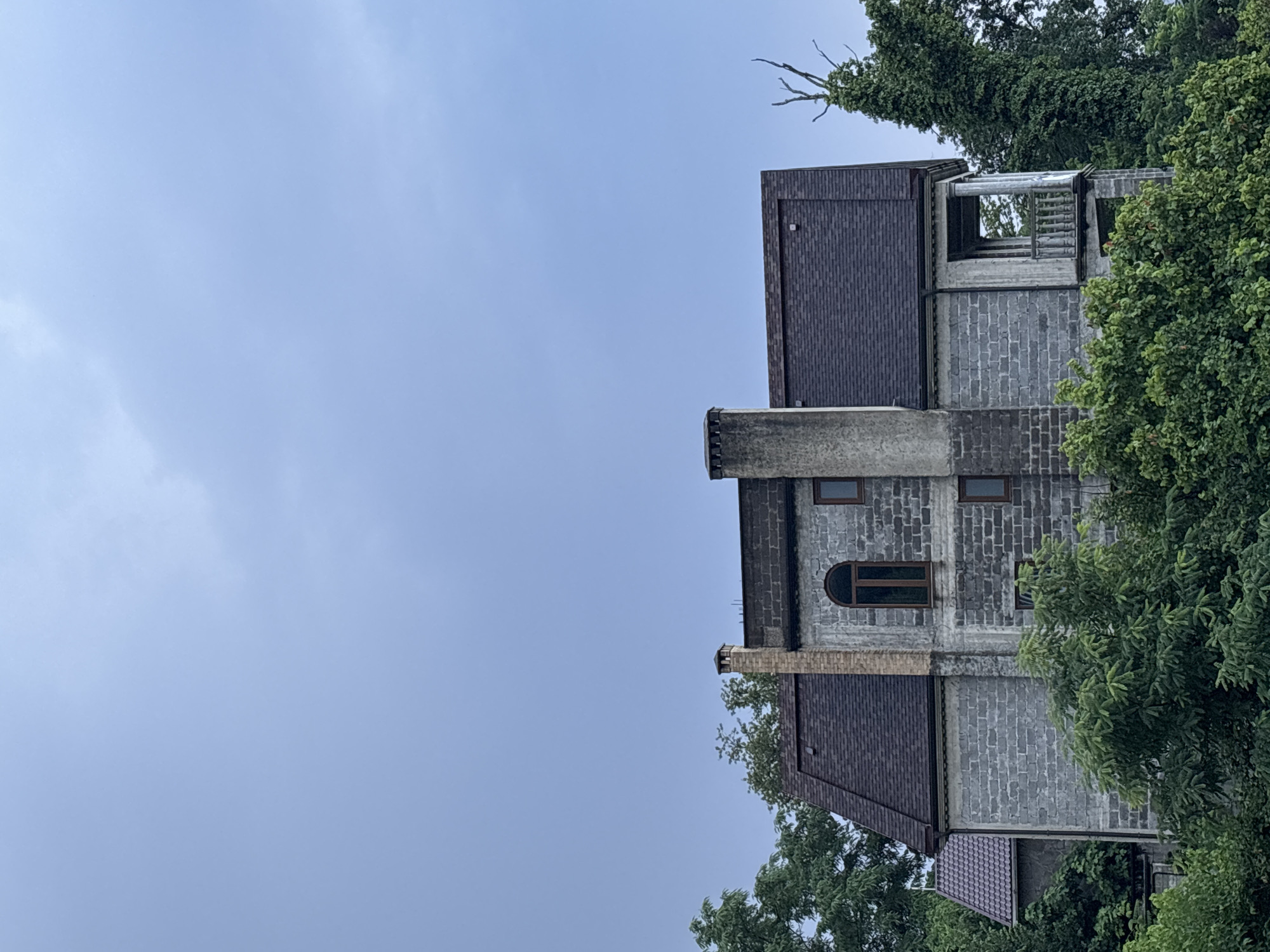 The image shows an unfinished, two-story building with a gray brick facade, surrounded by lush green trees. The building has a dark-shingled roof and features several windows, including one arched window. Two chimneys are visible on the roof, one made of brick and the other of concrete. A small balcony with decorative railings is also present on the right side of the building. The sky is overcast with a pale blue hue. The building is situated on a hillside, with dense foliage obscuring the lower portion. There are no people or animals visible in the image. The scene appears to be in a rural or suburban area, possibly on the outskirts of Sochi, Russia, given the subtropical vegetation and the style of the unfinished construction.