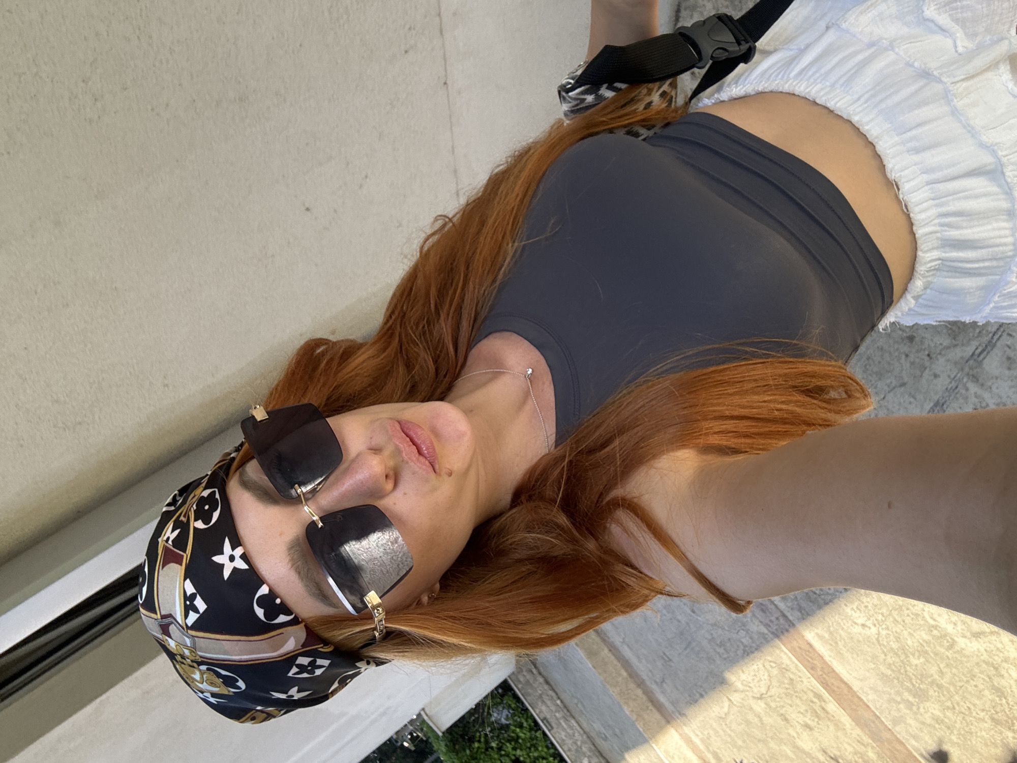 The media file shows a person lying on their back, with their head tilted to the side. The person has long, auburn hair and is wearing a dark grey tank top, white ruffled shorts, and a black and white patterned bandana on their head. They are also wearing large, square-shaped sunglasses with gold accents. A silver necklace with a small pendant is visible around their neck. A black strap with a buckle is visible around their waist, potentially part of a bag or accessory.
The scene appears to be outdoors, with a textured, light-colored wall behind the subject. The ground appears to be made of concrete or stone, with visible lines indicating paving. Sunlight casts shadows on the ground, suggesting it is daytime. The person's expression is relaxed, with their eyes closed and lips slightly parted. The overall mood is casual and summery.
The location is identified as Budva, Montenegro, which might be suggested by the outdoor setting, the style of architecture, or the presence of vegetation visible in the background, although specific landmarks are not clearly discernible. The time of day is likely afternoon due to the strong sunlight and shadows. The weather appears to be warm and sunny.
There is no visible text in the media file.