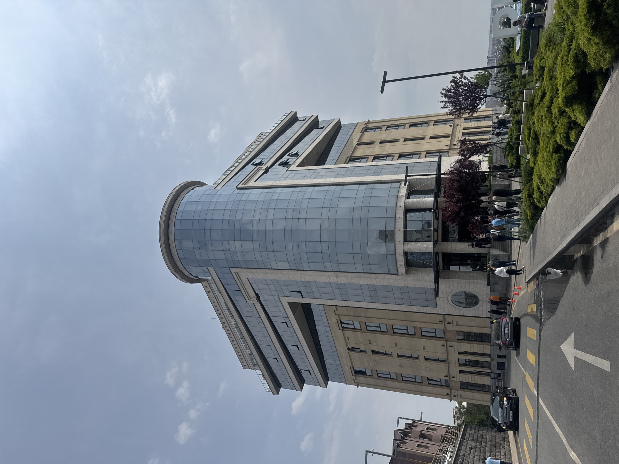A ground-level, long shot captures a modern glass and concrete building in Yerevan, Armenia. The building features a prominent cylindrical glass tower integrated with layered rectangular sections. The sky above is overcast with light, scattered clouds.
Adjacent to the building is a landscaped area with green bushes and small, red-leaved trees. A sidewalk runs alongside the street, where several people are standing or walking.
A paved road is visible in the foreground, marked with a large white arrow indicating the direction of traffic flow. Two cars are parked along the street, a black sedan and a gray SUV.