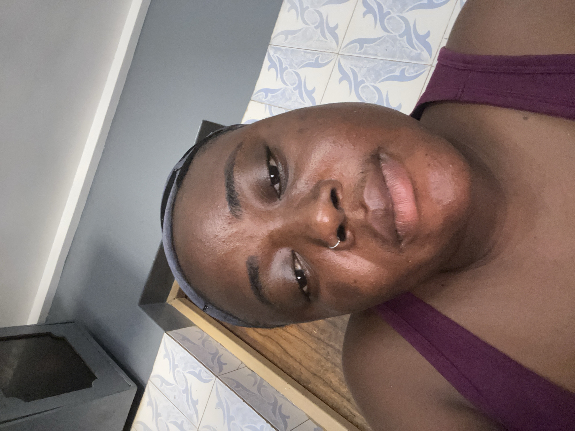 A selfie of a dark-skinned woman with a nose ring and a gray headband wearing a purple tank top. The background includes a cabinet, gray and white walls, and patterned tiles. There is no information to confirm if the photo was taken in the city of Jos, Nigeria.