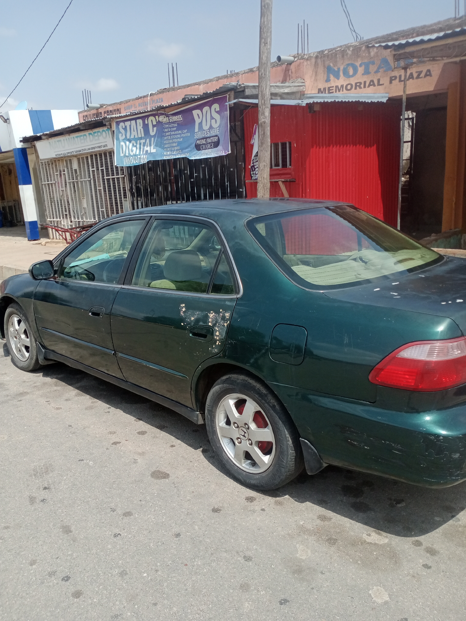 The image shows a dark green sedan parked on a paved road in Jalingo, Nigeria. The car is positioned at an angle, with its rear facing towards the right side of the frame. The vehicle exhibits signs of wear and tear, including scuffs and peeling paint on the rear passenger door and fender. The car's wheels are visible, with one on the left side showing a silver alloy rim and red brake calipers. The background reveals a street scene with buildings and signage. To the left, a building with blue and white striped columns and barred windows is visible, with a sign reading "NATU WATER DEPOT". Next to it, a banner with "STAR 'C' DIGITAL INNOVATION" is displayed. Further to the right, a red corrugated metal building has a sign that reads "NOTA MEMORIAL PLAZA". The sky above is clear and blue. The road surface shows patches of discoloration, possibly oil stains.