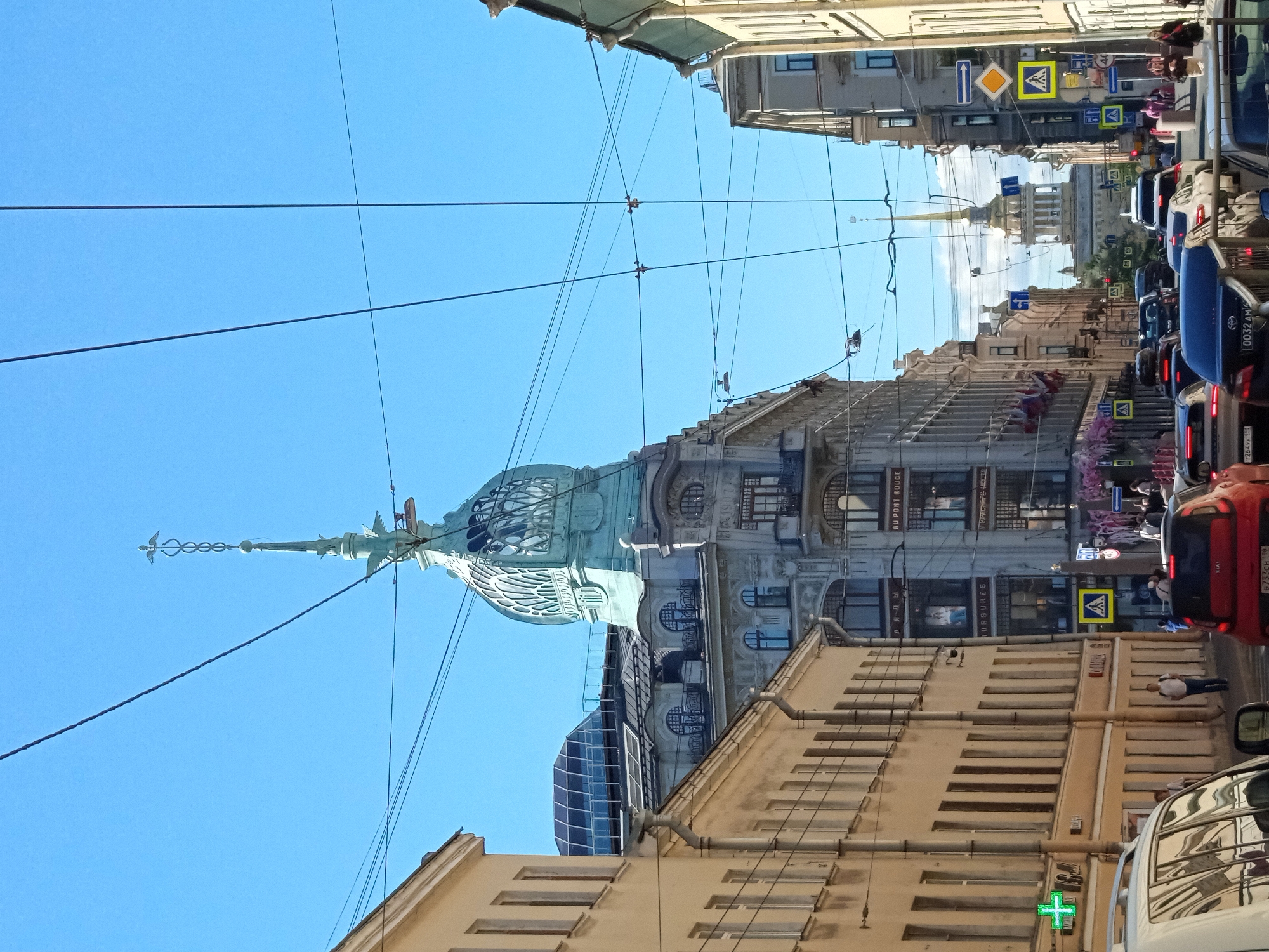 The image depicts a street scene in Saint Petersburg, Russia, likely on a clear, sunny day given the bright blue sky. The perspective is from street level, looking upwards and slightly down the street. The orientation of the photo is unusual, with the scene appearing sideways.
Dominating the mid-ground is a distinctive building with an ornate, green-domed spire topped by a golden finial. The architecture suggests a historical European city, with ornate facades, detailed window frames, and decorative elements on the buildings. Several other buildings line the street, some appearing older and classical in style, while others are more modern, including one with a large, curved glass facade.
The street is lined with cars, indicating it is a busy urban area. Traffic signals and pedestrian crossing signs are visible, suggesting an active intersection. Overhead, a complex network of electrical wires crisscrosses the sky, a common sight in many cities.
There are a few people visible, mostly appearing as small figures in the distance, either walking on the sidewalks or inside cars. One person is standing on a sidewalk near a building.
Some text is partially visible on the buildings, including what appears to be shop signs in Russian, such as "AU PONT ROUGE" which is French, suggesting a historical or cultural influence. A green cross symbol, often indicating a pharmacy, is visible on one of the lower buildings. The overall mood of the image is that of a bustling city on a pleasant day, with a focus on the architectural grandeur and the urban environment.