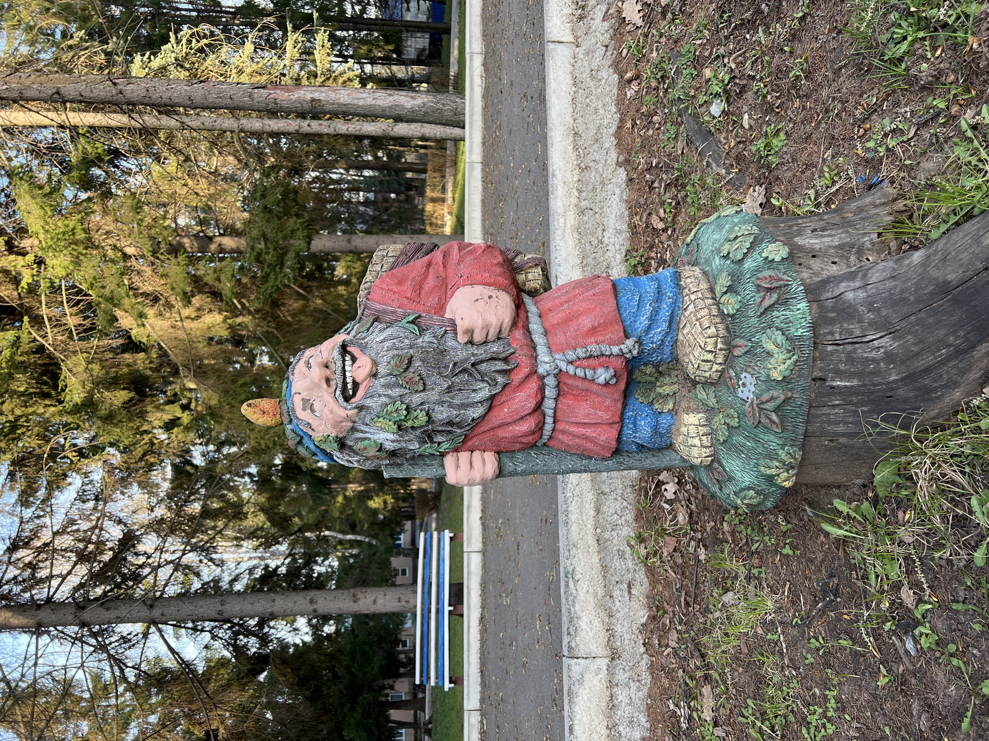 The image depicts a colorful garden gnome statue, seemingly made of painted concrete or similar material, standing next to a sidewalk in an outdoor environment. The gnome is positioned next to the trunk of a tree, with some foliage and ground cover visible around the base of the tree. The background includes trees and a partially visible bench with blue and white slats. A paved sidewalk extends into the distance, suggesting a park-like or recreational area. The location is Sanatoriya Yumatovo Imeni 15-Letiya Bassr, Russia.