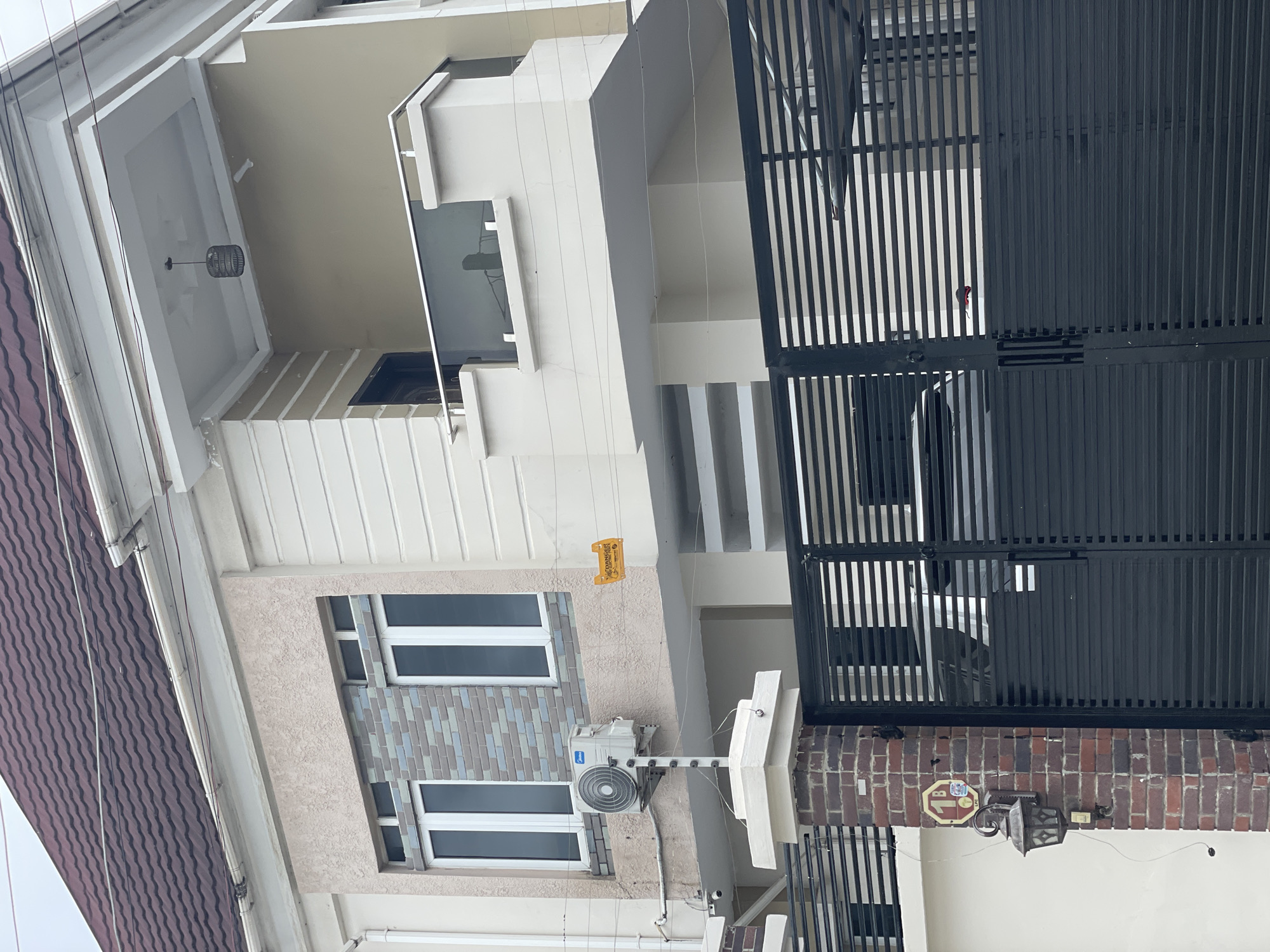 This image depicts the exterior of a modern, multi-story building in Ado, Nigeria. The building features a dark red tiled roof, cream-colored exterior walls with some textured and striped sections, and multiple windows. An air conditioning unit is mounted on the exterior wall. A black metal gate is visible in the foreground, partially obscuring a paved area and potentially vehicles. A brick pillar with a lantern and a circular yellow sign is positioned next to the gate. The environment appears to be a residential or commercial area.