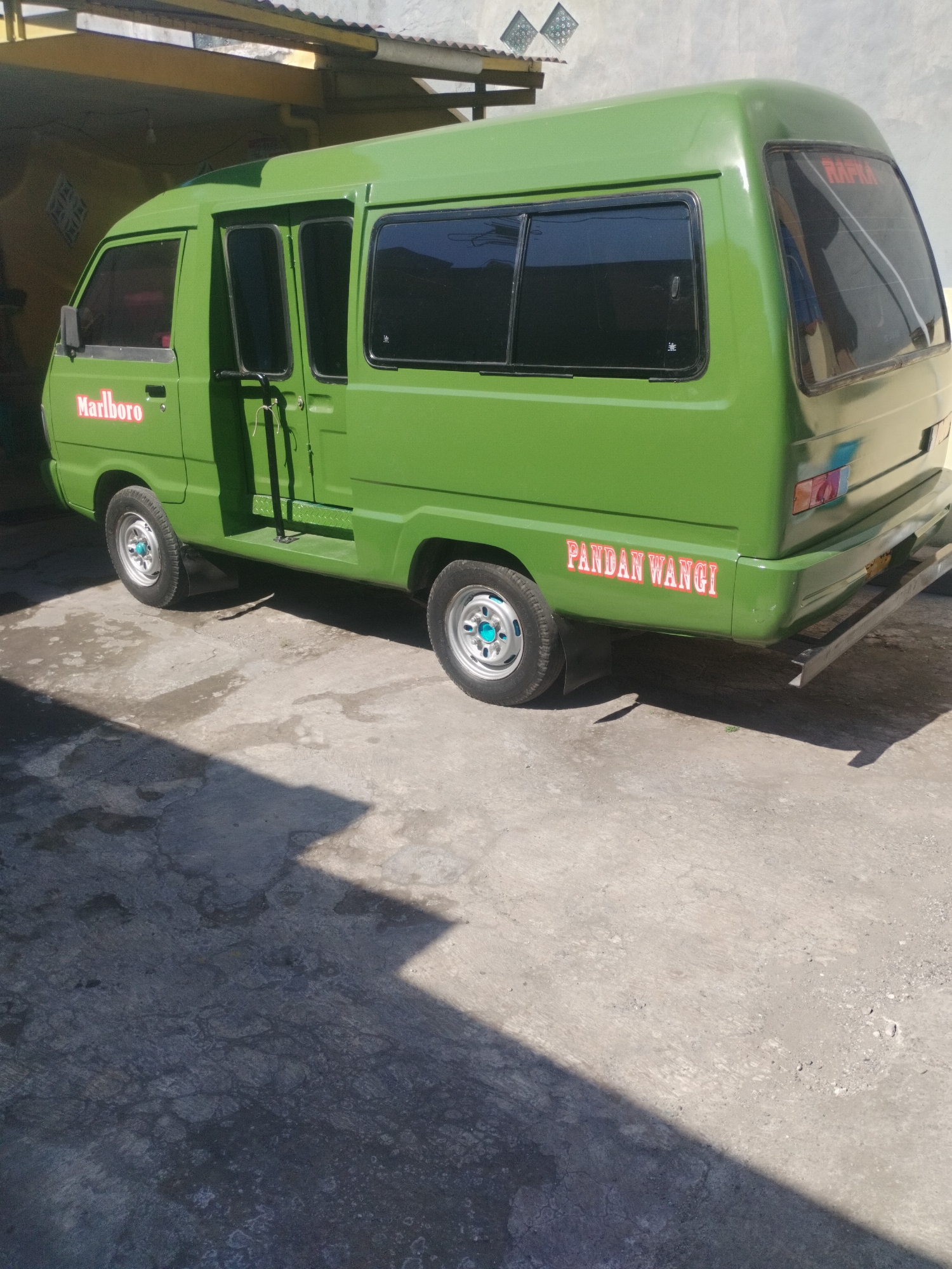 A bright green minibus is parked on a concrete surface, casting a shadow. The minibus has tinted windows and logos on its sides. The logo on the left reads "Marlboro" in red lettering, and the logo on the right reads "PANDAN WANGI" also in red lettering. The minibus appears to be a passenger vehicle, possibly a public transport vehicle. The environment is somewhat industrial, with a building structure behind the vehicle that appears to be a covered parking area or workshop. The overall scene is well-lit, suggesting it is daytime. Based on the style of the minibus and the signage, the image likely depicts a scene in Surabaya, Indonesia.