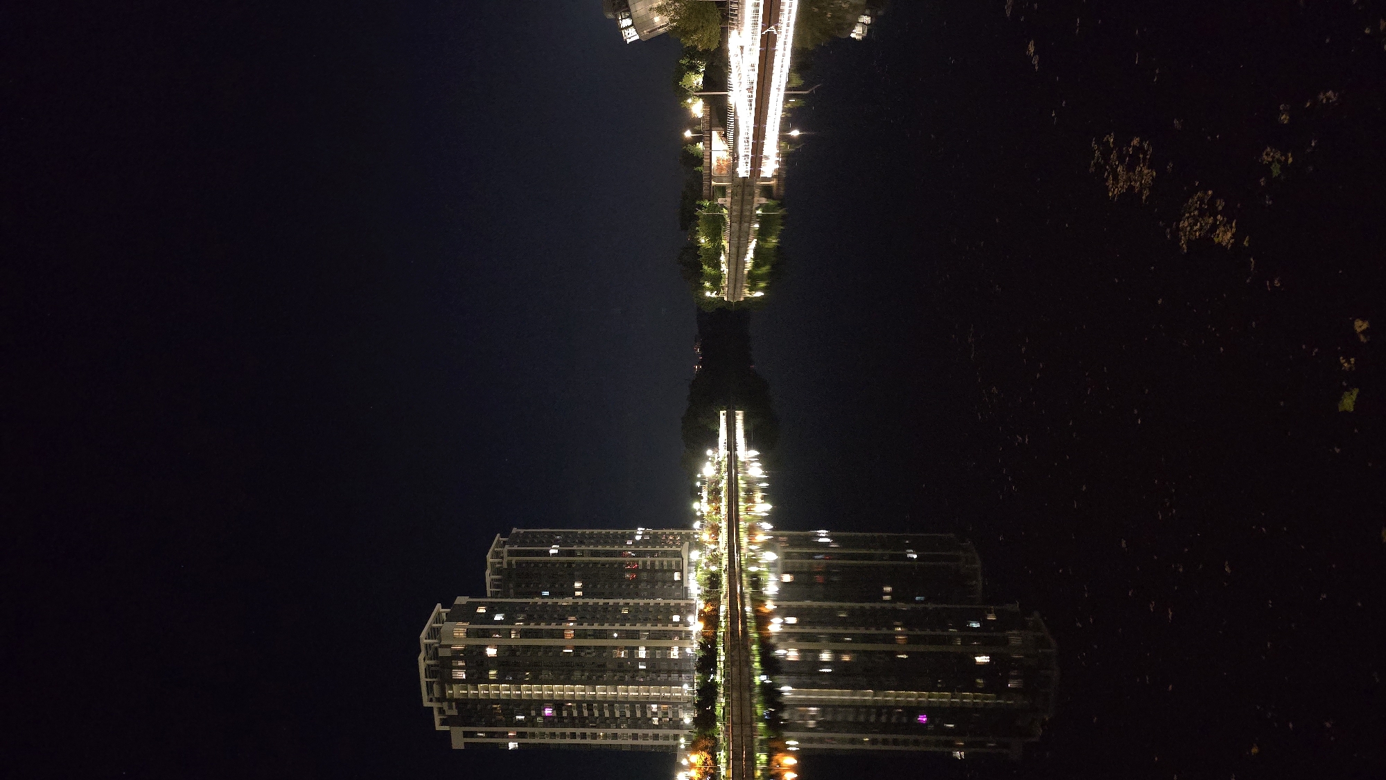 A nighttime, vertical, medium-shot photograph captures the reflection of illuminated high-rise buildings and city lights in the dark water of the Dnipro River in Kyiv, Ukraine. The two prominent buildings on the left are tall residential towers with many windows lit from within, their lights creating a vibrant, colorful reflection on the water's surface. A shoreline with trees and streetlights is visible between the buildings and the water. The right side of the frame shows a more distant, lower building with illuminated windows and lights along its waterfront. The dark sky above is clear and shows no celestial bodies. The overall scene is one of urban tranquility at night, with the city's lights creating a beautiful contrast against the dark water and sky.