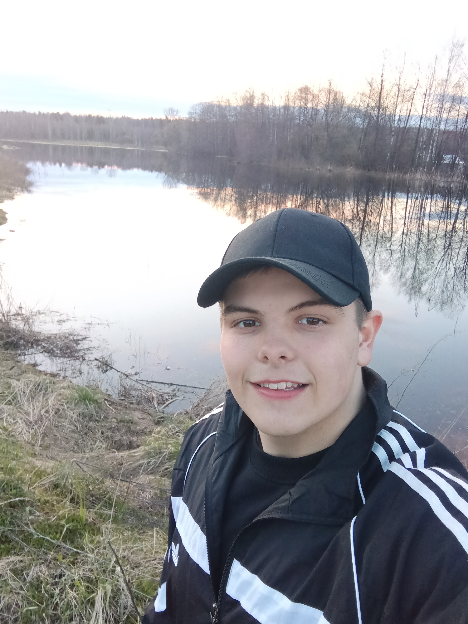 A young male, wearing a black baseball cap and a black track jacket with white stripes, stands in a selfie pose. He is positioned on a riverbank with dry grass. In the background, a lake reflects the sky and bare trees. The scene is located in Красный Маяк, Russia.