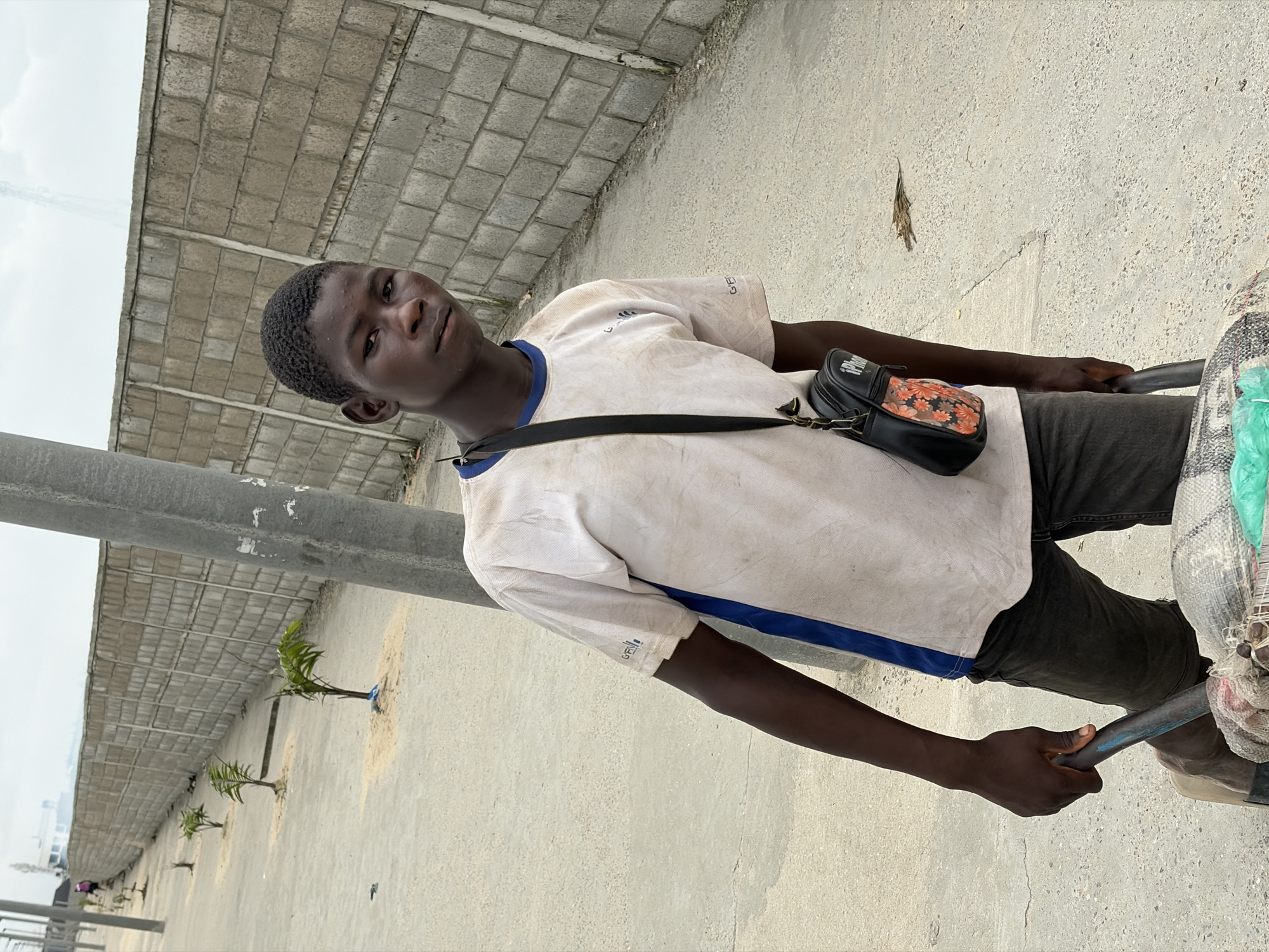 A young Black male stands outdoors, holding the handles of a cart filled with wrapped goods. He is wearing a dirty white t-shirt with blue trim and dark pants. A black bag with a floral pattern is slung across his chest. The background shows a concrete wall and a sidewalk lined with young palm trees. The scene appears to be in Igboefon, Nigeria.
