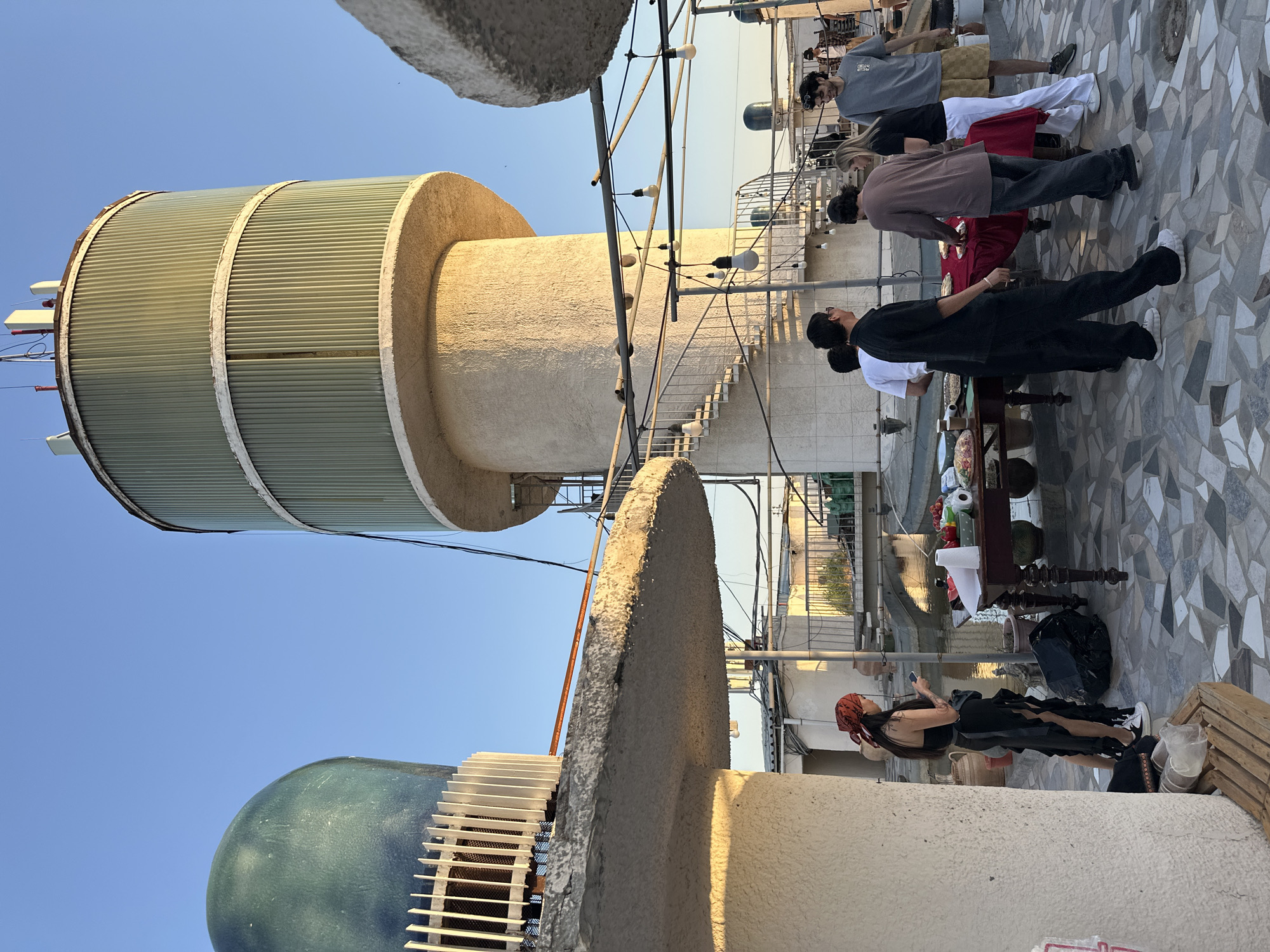 This image shows a rooftop gathering in Tashkent, Uzbekistan. A group of people are standing around a table laden with food and drinks. They appear to be interacting and socializing. The backdrop features urban architecture, including distinctive cylindrical structures: one with a corrugated metal exterior and another with a blue dome and white grating. String lights are strung overhead. The ground is covered with a patterned tile. The sky is clear and blue. A woman in a black dress and red headscarf stands apart, looking at her phone.