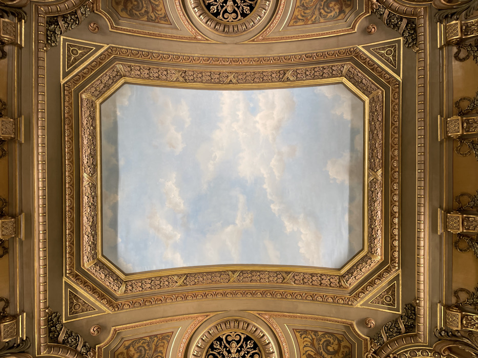An ornate ceiling features a large, rectangular central panel painted with a blue sky and scattered white clouds. The panel is framed by multiple layers of decorative molding with geometric patterns and gold accents. The surrounding ceiling area has similar ornate detailing, including circular medallions with intricate black filigree. The color palette is dominated by gold, beige, and off-white, creating a rich and luxurious aesthetic.