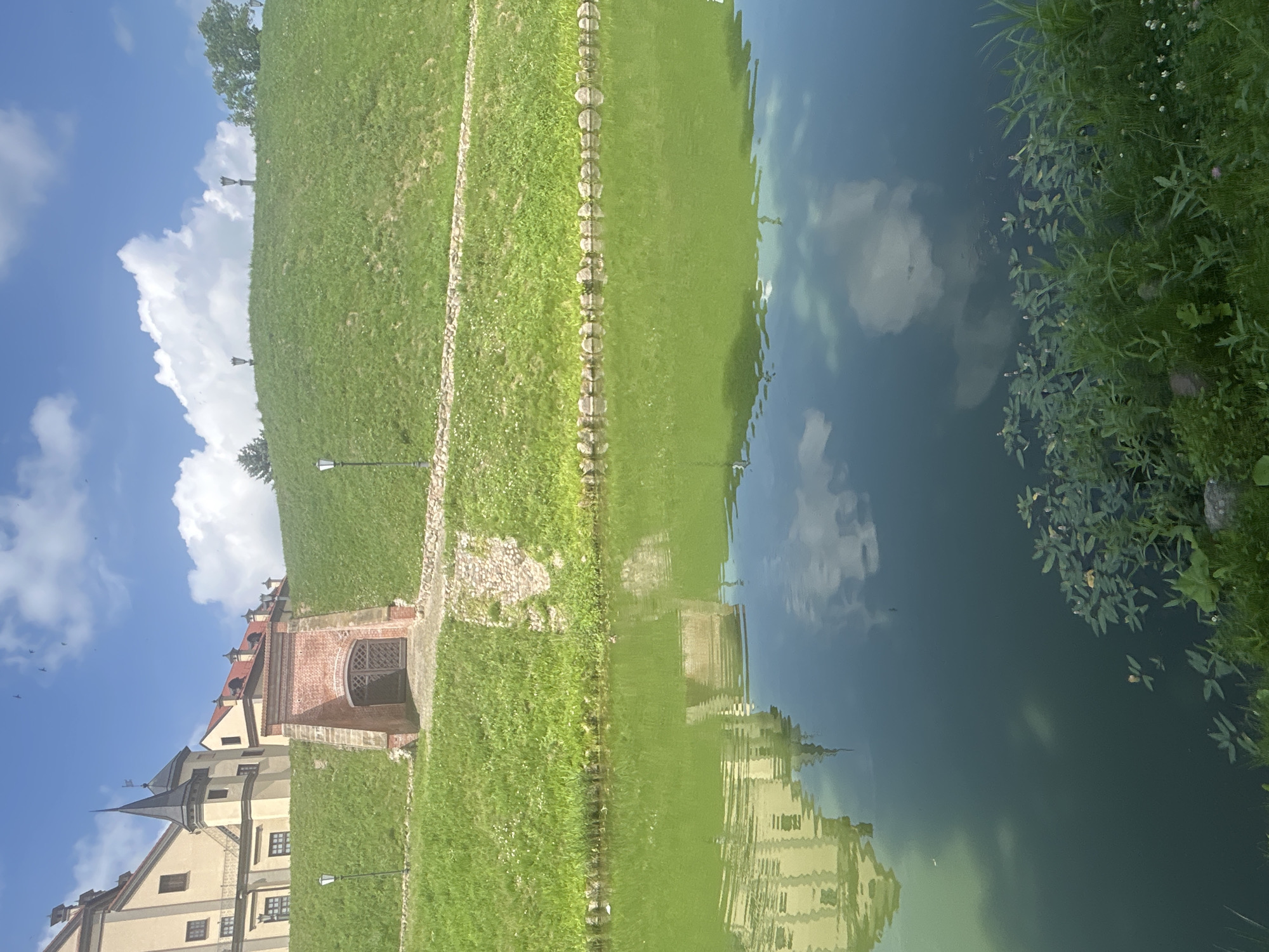 The image depicts the Nesvizh Castle in Nesvizh, Belarus, on a sunny day with scattered clouds. In the foreground, a tranquil body of water reflects the castle's architecture and the sky. The water is a vibrant green hue, with lily pads and reeds growing along its edge.
Dominating the midground is a steep, grassy embankment that leads up to the castle. A brick-built structure with an arched wooden gate is partially embedded into the hillside, serving as an entrance. The castle itself, a grand, pale yellow building with dark spires and red-tiled roofs, is situated atop the embankment. Several lampposts are visible along the slope.
The sky above is a bright blue, punctuated by fluffy white clouds. The overall scene is serene and picturesque, showcasing the historical grandeur of the Nesvizh Castle and its surrounding landscape. There are no people or animals visible in the image.