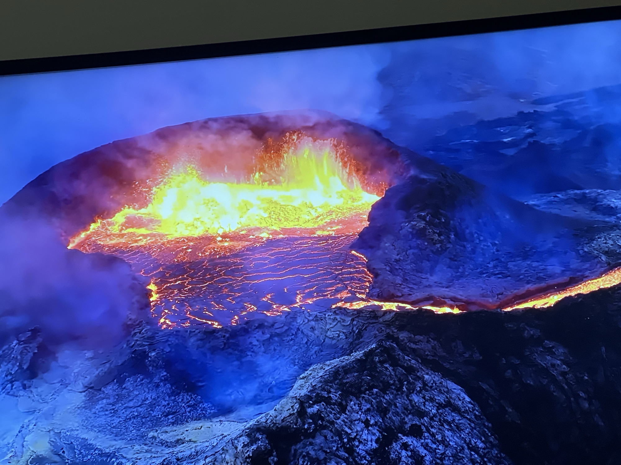 This is a close-up, high-angle shot of a volcanic eruption. Molten lava, glowing bright yellow and orange, fills the caldera of the volcano, with some spewing out over the rim. Thick plumes of smoke and ash, colored purple and red from the heat, rise from the crater. The surrounding landscape is dark and rocky, rendered in deep blues and purples, suggesting a night scene or the atmospheric effects of the eruption. The image appears to be a photograph of a screen displaying this natural phenomenon, indicated by the presence of a dark bezel at the top of the frame. There is no information about Sofia, Bulgaria in relation to this image.