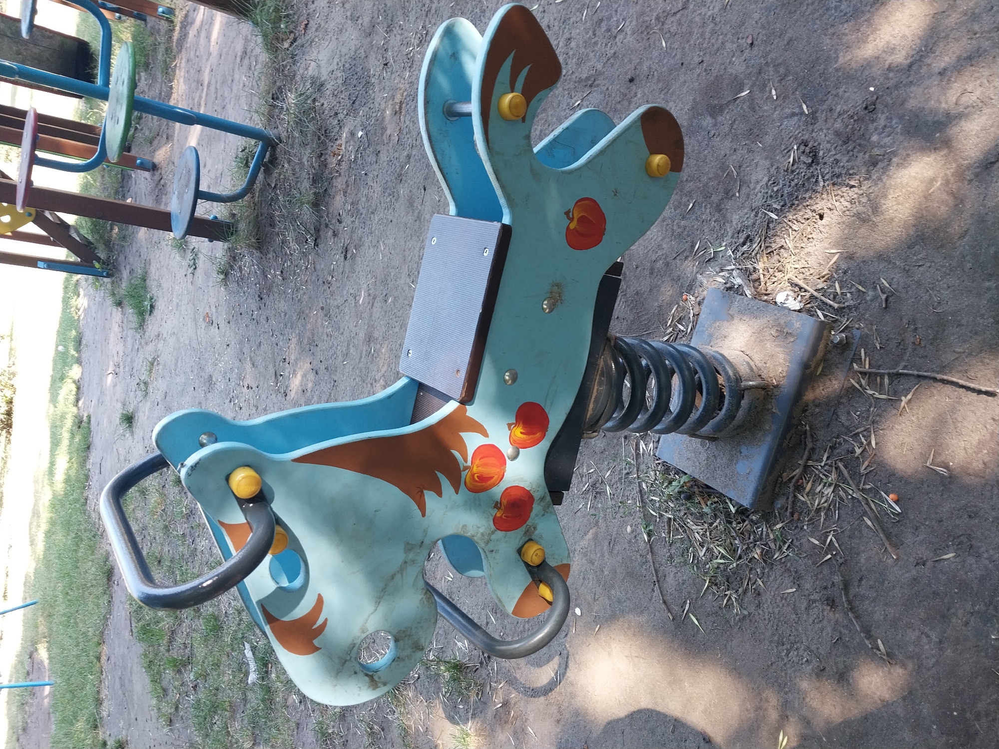 A blue spring rider shaped like a horse is located on a dirt playground in Doslidne, Ukraine. The horse has brown mane and tail details and red apple illustrations on its sides. Yellow caps are visible on its ears, eyes, and rear. A dark grey handlebar arches over the horse's neck, and a black seat is positioned on its back. The horse is mounted on a black spring attached to a grey metal base. The playground ground is covered with dirt, scattered leaves, and small debris. In the background, other playground equipment, including a swing set, is partially visible. The lighting suggests it is daytime, with sunlight casting shadows on the ground.