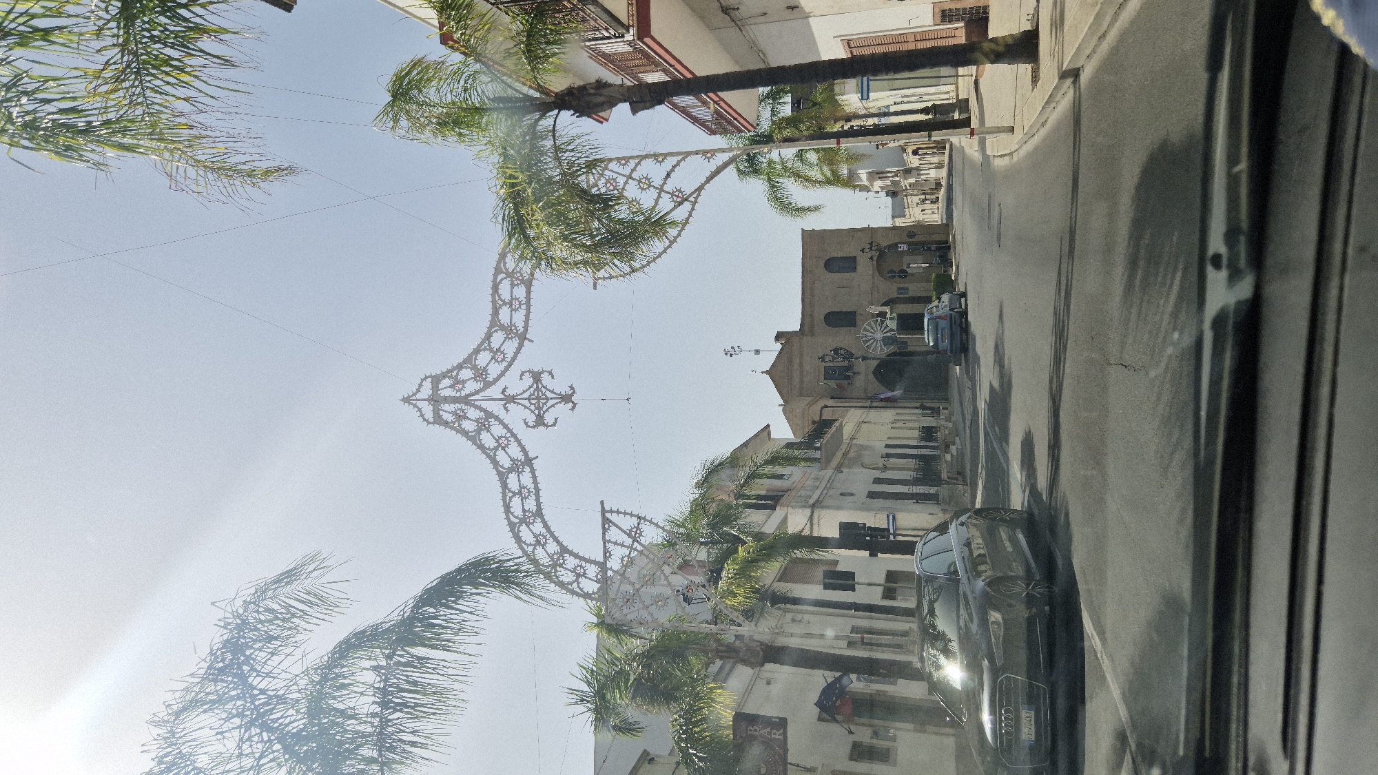 The image shows a street in Maruggio, Italy, with buildings on either side and palm trees. There is a decorative archway spanning the street, adorned with lights and intricate patterns. A dark-colored Audi A6 Avant is parked on the left side of the road, facing forward. In the distance, a light blue car is visible. The sky is clear and bright, with the sun casting a glare on the scene. The architecture suggests a Mediterranean setting, and the overall atmosphere is that of a sunny day in a town square or a main thoroughfare.