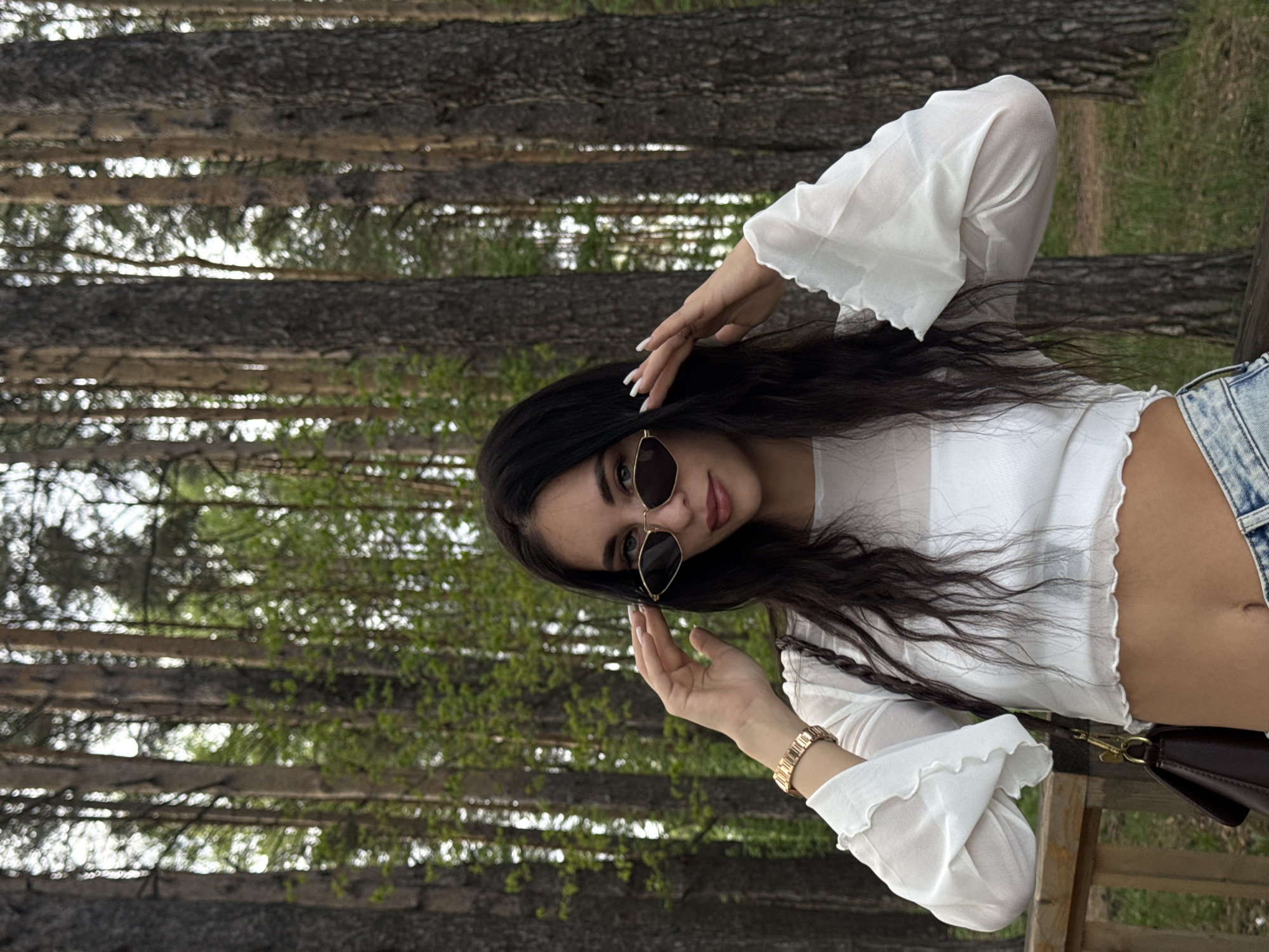 A woman with long dark hair and sunglasses poses outdoors in a wooded area. She is wearing a white, semi-sheer blouse with ruffled sleeves and light-wash denim shorts. The background consists of a dense forest with tall trees, some showing early signs of spring foliage. The location appears to be Chaikovsky, Russia. She is standing next to a wooden fence. A brown purse hangs on her shoulder, and she is wearing a gold watch. She is holding her left hand up near her sunglasses. Her nails are painted white.