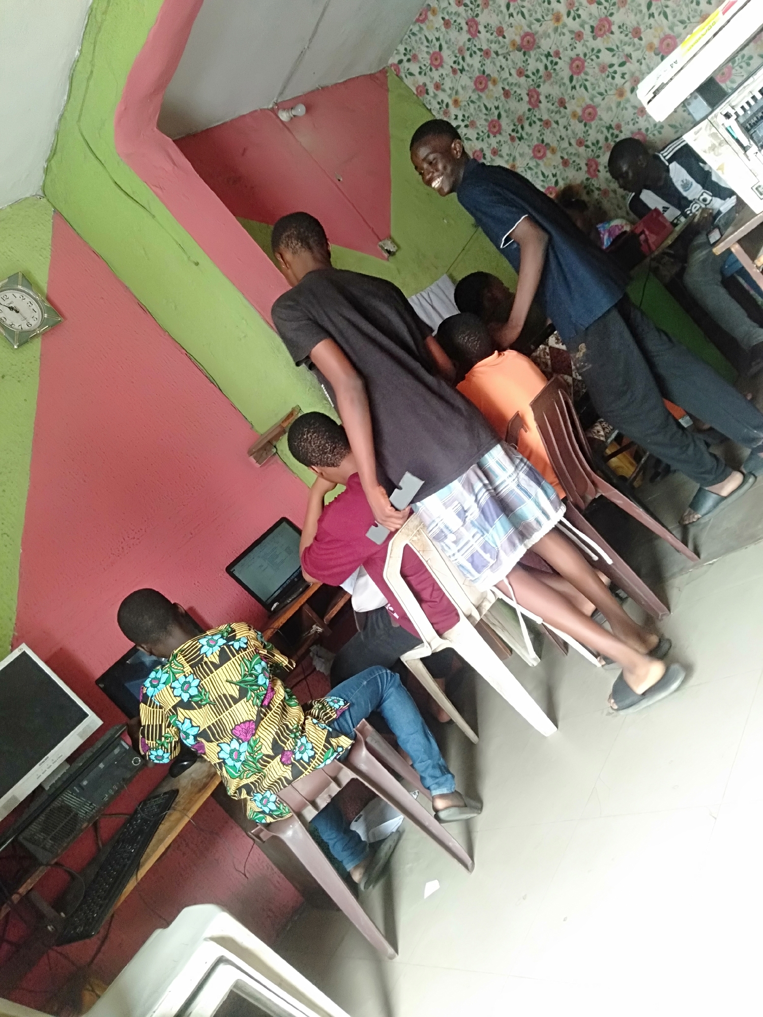 This image depicts an indoor scene within what appears to be an internet café or computer lab in Shangisha, Nigeria. The room is brightly decorated with walls painted in striking sections of pink and lime green, contrasting with a floral-patterned wallpaper on the back wall. The floor is tiled.
The primary subjects are approximately seven young males, likely teenagers or young adults, engaged in using desktop computers. Most are seated on plastic chairs and focused on their individual screens. On the left, a young man in a vibrant yellow, blue, and black patterned shirt is seen operating a computer. In the center, another individual wearing a maroon shirt is also at a computer. A young man in a black t-shirt and plaid shorts stands and leans over a chair, looking at a screen, while another in an orange t-shirt is partially visible behind him. Towards the right, a young man in a dark blue polo shirt stands smiling broadly, looking towards the left, contributing a cheerful mood to the scene. Further right, another person in a black and white striped jersey is seated at a table, seemingly using a handheld device.
Notable details include several desktop computer setups with monitors, keyboards, and CPU towers. A small, square, silver-framed wall clock on the left wall indicates the time is approximately 1:55. Various plastic chairs, both white and brown/maroon, are used for seating. Electronic equipment and shelving are visible on the far right. The activity suggests a communal space for digital engagement. No legible text is visible apart from the Roman numerals on the clock.
