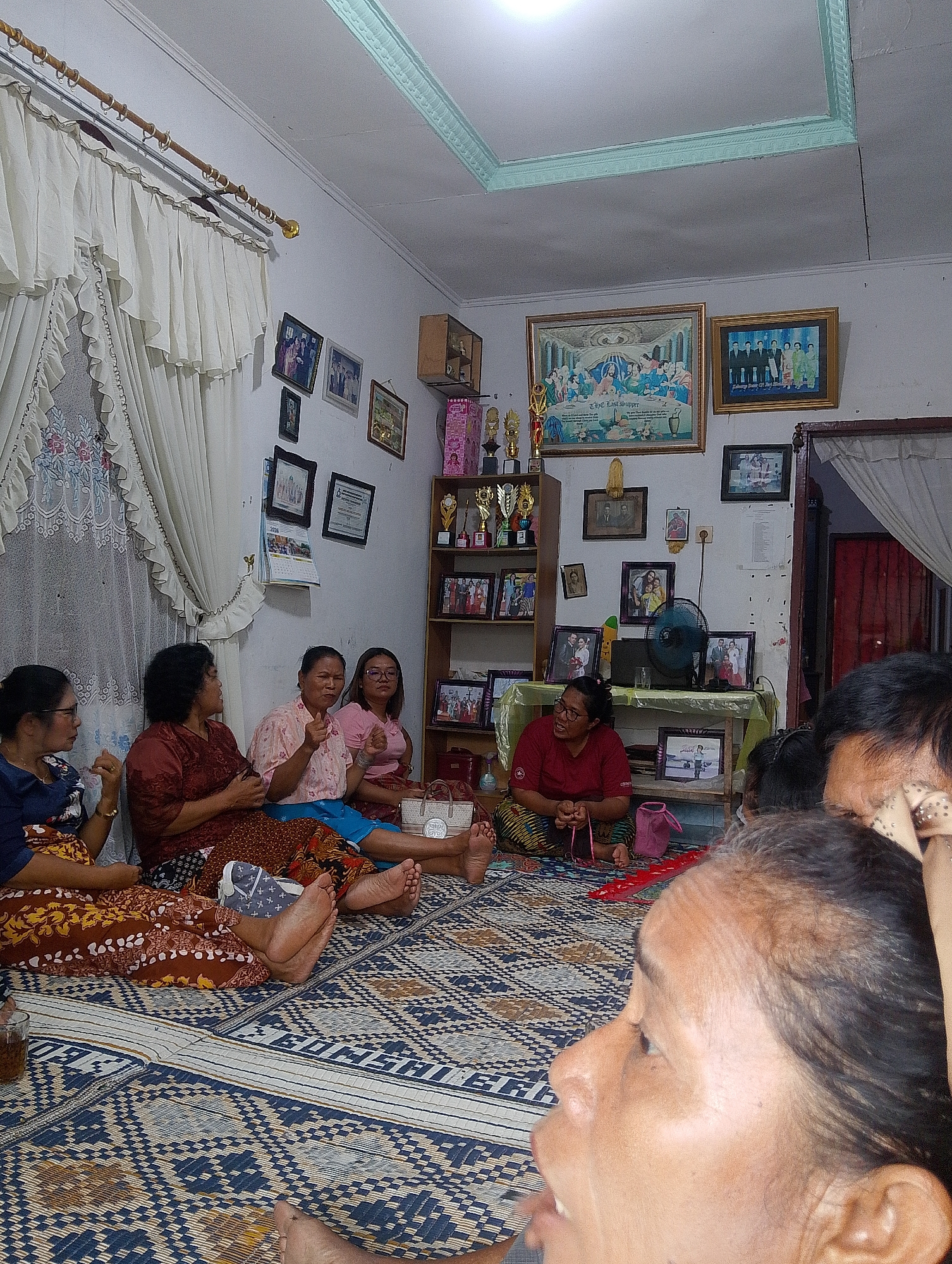 The image shows a group of women sitting on woven mats in a room. The room appears to be a living area, with white walls, a patterned floor covering, and a decorative ceiling. There are several framed pictures on the wall, including what appears to be a depiction of "The Last Supper" and a family portrait. A shelf unit on the right side of the room holds trophies and more framed photographs. A fan is visible on a table, along with a glass and a pink bag. Curtains hang on the left side of the room. The women are dressed in traditional clothing. One woman in the foreground is looking away from the camera, and her face is the most detailed in the image. The lighting suggests it is daytime, possibly with artificial light supplementing natural light from an unseen source. The scene appears to be a social gathering. The location is identified as Naga Jaya 1, Indonesia.