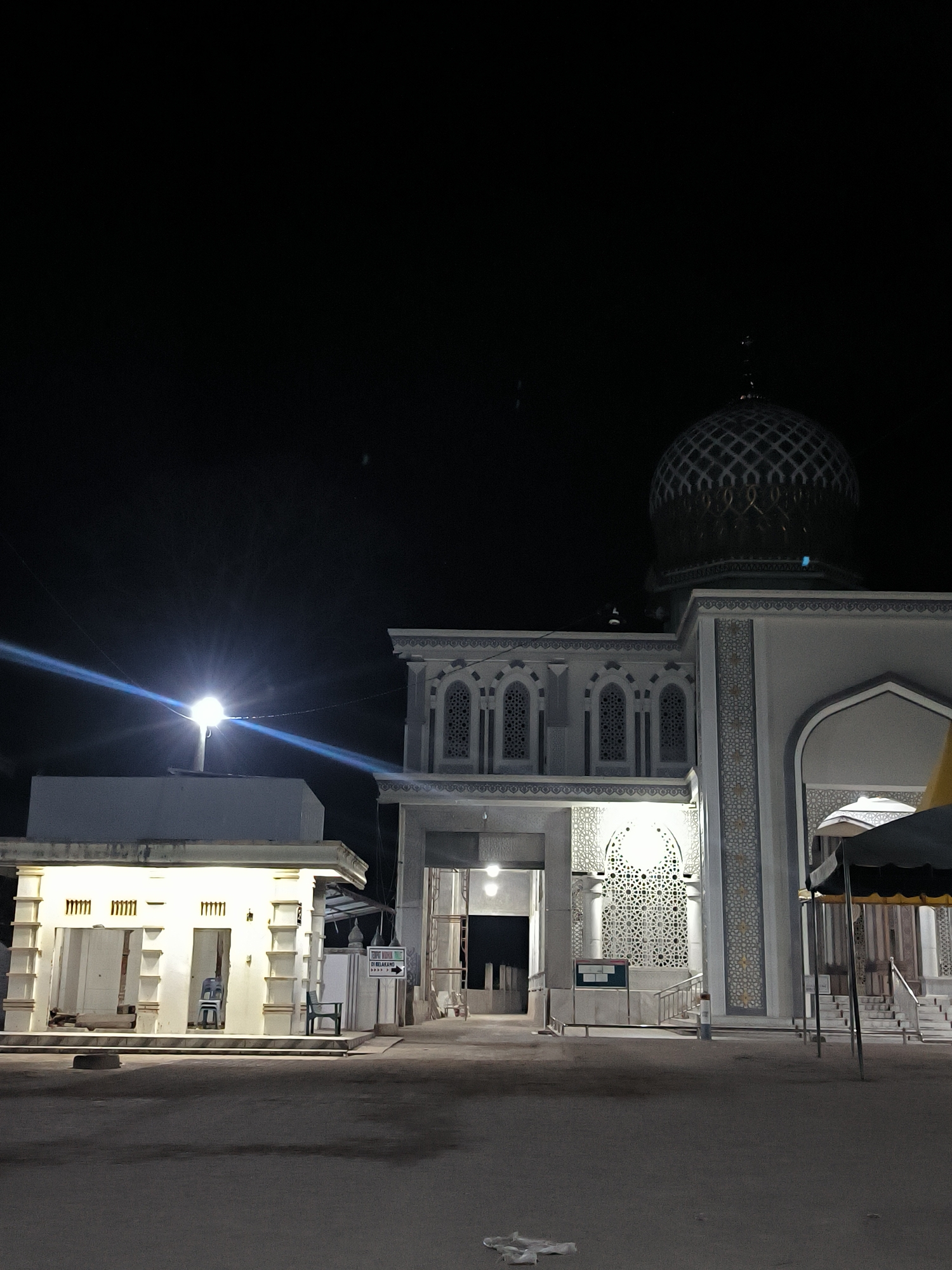 At night, a brightly lit mosque with a patterned dome is visible in Keudealue Ie Puteh, Indonesia. An adjacent smaller building with open stalls and a solitary chair is also illuminated. A sign is present near the mosque's entrance. The ground is dark and appears to be a paved area. The overall environment is dark.