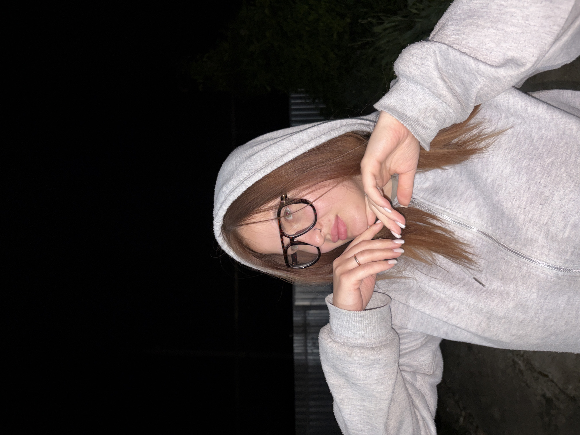 A young woman wearing a gray hooded sweatshirt and glasses is posing for a photo. She is standing outdoors at night. Her hands are near her face. The location is possibly in or near Novopokrovskaya, Russia. In the background, there is some foliage and what may be a fence.