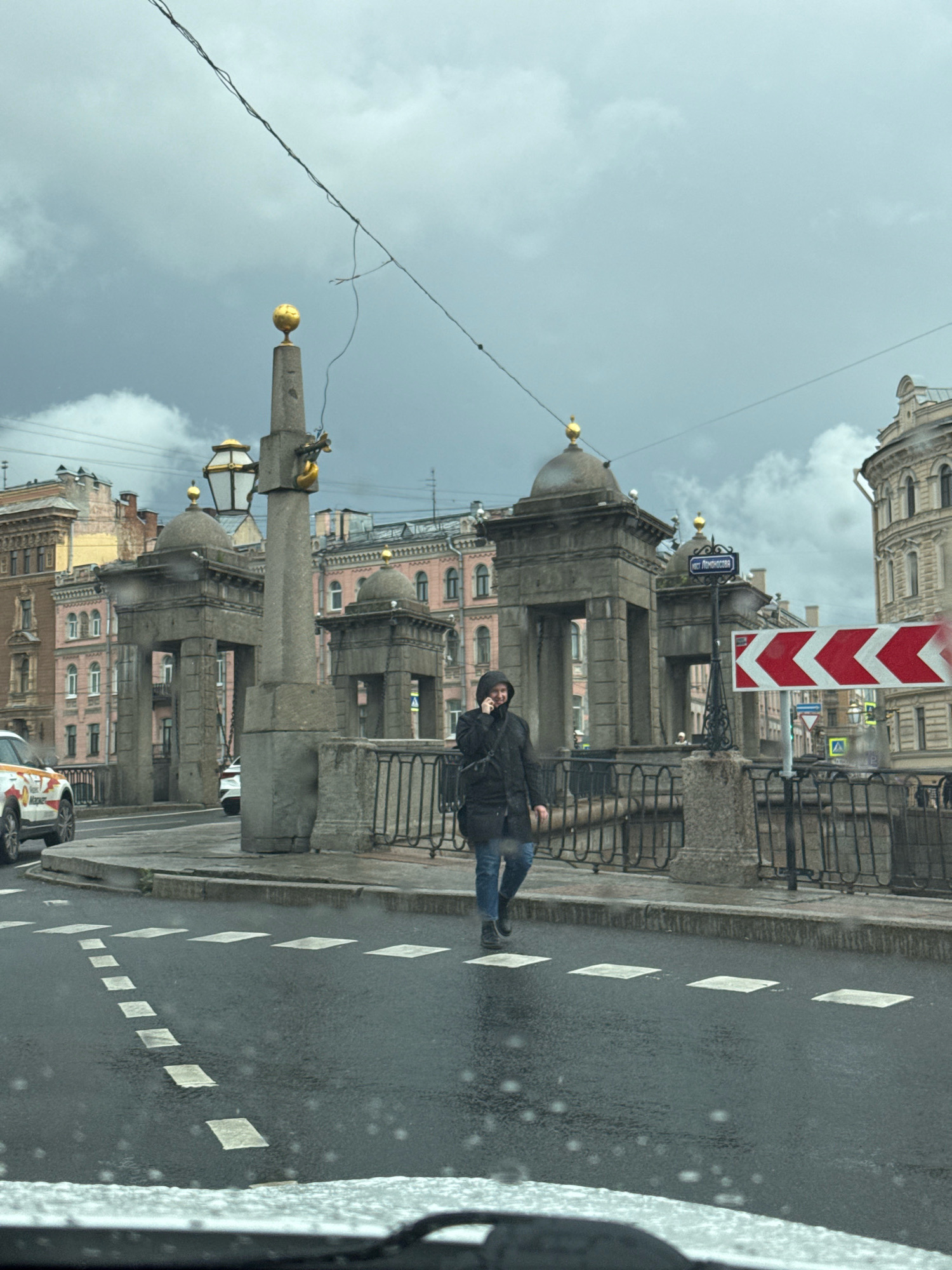 The image shows a person in a black hooded coat walking across a wet street in Saint Petersburg, Russia. The person is on the phone and appears to be crossing a pedestrian crossing. In the background, there are architectural structures that are characteristic of Saint Petersburg, including stone columns topped with golden spheres and domes. There is also a road sign visible. The sky is overcast and cloudy, and there are visible raindrops on the road and on what appears to be the windshield of a vehicle in the foreground, suggesting it might be raining or has recently rained. A white car with orange and yellow markings is parked on the left side of the street. The overall scene depicts an urban environment with historical architecture under a gloomy sky.