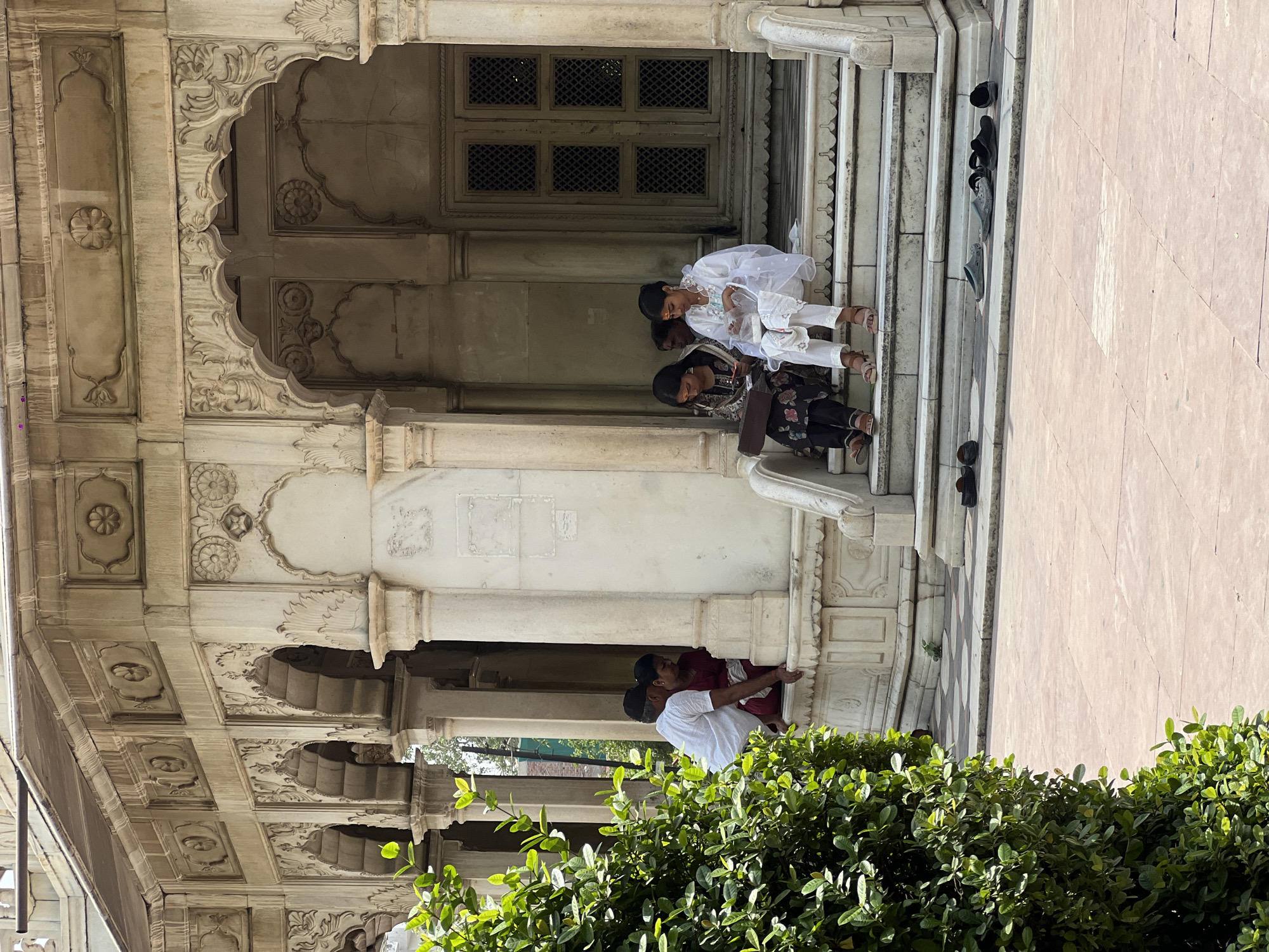 The image depicts people sitting on steps adjacent to an ornate architectural structure, likely in Varanasi, India.
**Environment:** The architecture is characterized by light-colored stone, possibly marble, with intricate carvings on the facade and arches. The environment appears to be a courtyard or outdoor space with lush green bushes and foliage in the foreground, partially obscuring the lower portion of the building. The steps are light-colored and several pairs of shoes are scattered on the steps above the people.
**People:** Several people are observed on the steps. Two children, dressed in white, are seated at the top of the steps. Below them, a woman dressed in dark patterned clothing is sitting. Further back and below, two people are standing and embracing.
**Actions:** The people are seated on the steps and possibly interacting with each other. The others further back and below are embracing.
**Overall:** The scene captures a casual moment in a historical setting, suggesting a blend of cultural heritage and everyday life in Varanasi. The juxtaposition of the intricate architecture with the relaxed posture of the people on the steps creates a contrast between permanence and transient human presence.