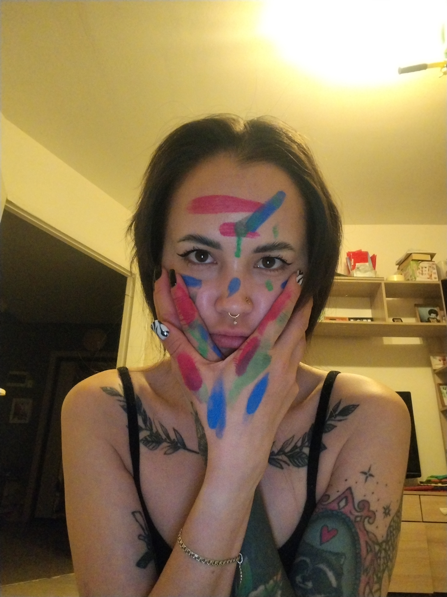 A woman is centered in the frame, her face marked with strokes of red, green, and blue paint. Her hands, also covered in the same paint, are pressed against her cheeks. She has dark hair, black eyeliner, and a nose piercing. She wears a black tank top.
Tattoos are visible on her chest and arms.
The background reveals an interior setting. A doorway is to the left, showing another room. To the right, a shelving unit with various items sits against the wall, next to a television and a dresser. The location is identified as Noviy, Russia.