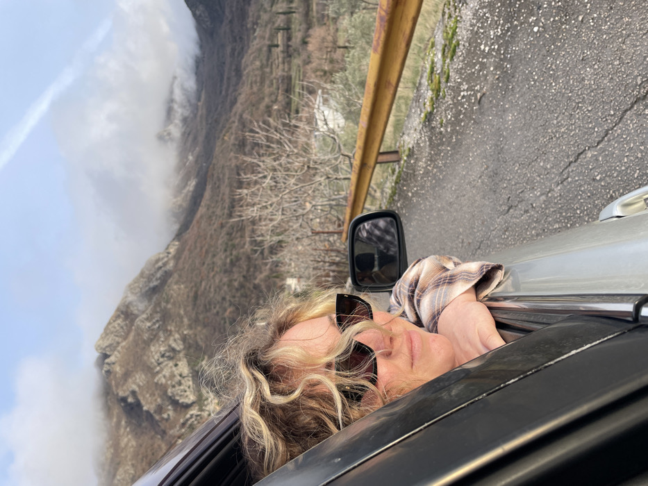 The image, oriented sideways, captures a person leaning out of the passenger window of a silver vehicle against a mountainous backdrop in Zupci, Montenegro. The subject, a person with light, wavy hair, wears dark sunglasses and a plaid shirt, appearing to be relaxed with eyes closed or squinting as they enjoy the passing scenery. Their arm rests casually on the car's window frame.
The background features a dramatic mountain range, partially obscured by dense white clouds or fog that cling to the peaks and slopes. The mountains show sparse, wintry vegetation, with numerous bare trees. To the right of the vehicle, a paved road is visible, bordered by a rusty yellow guardrail, with some moss growing along its edge. The lighting suggests it is daytime, with bright yet diffused sunlight. No visible text is present in the image. The scene evokes a tranquil moment of travel through a picturesque, elevated landscape.