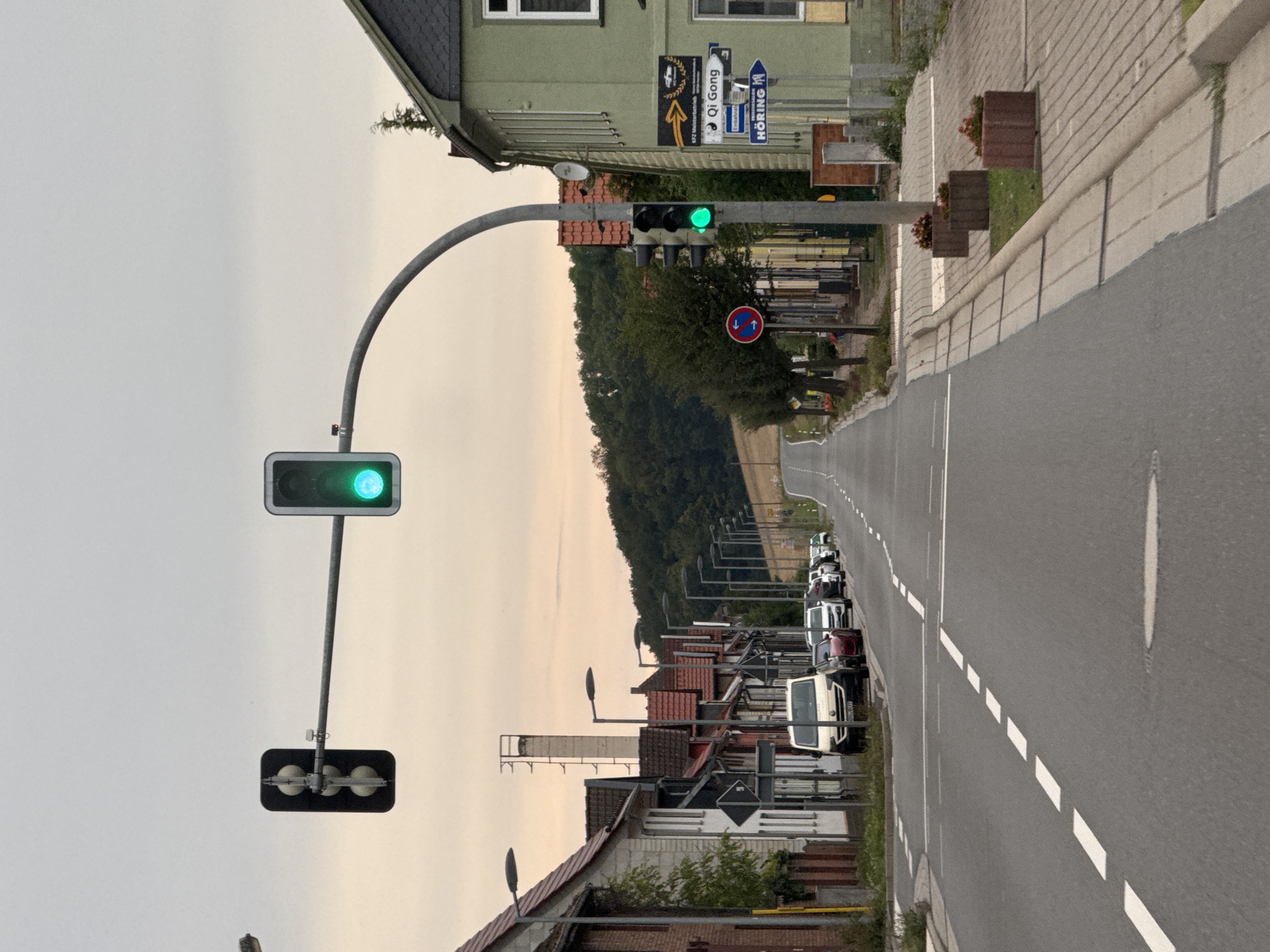 The image depicts a view of the street in Niedersachswerfen, Germany, during daylight hours with an overcast sky. The foreground is dominated by an asphalt road with white lane markings. On the right side of the road, a sidewalk is visible, lined with planters containing red flowers. Buildings with pitched roofs line the left side of the street, and the right side features a light green building with dark gray shingles. Traffic lights are prominently displayed, with the primary traffic light over the road showing a green light. Another traffic light on a pole on the right side also shows a green light. Various cars are parked along the side of the road on the left. In the distance, a forested hillside rises, and a road curves into the distance. A tall, industrial-looking structure, possibly a chimney, is visible behind the buildings on the left. Road signs are present, including a blue sign with white arrows indicating "Qi Gong" and "HÖRING". A circular red and blue sign with white arrows is also visible. No people are present in the frame.