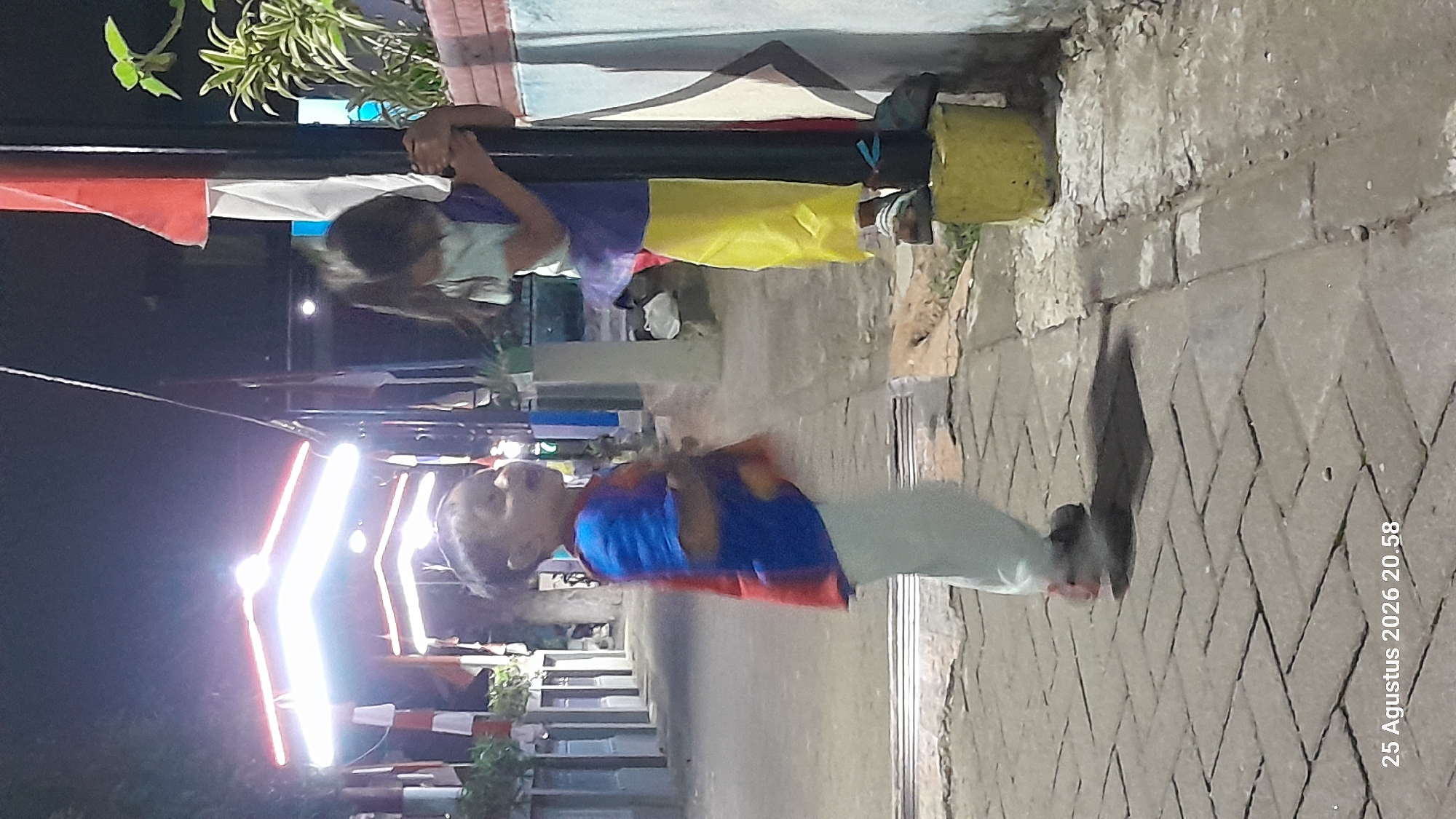Two young children are outdoors at night, with bright lights illuminating the scene from overhead. The child on the left, wearing a blue and red shirt and light-colored pants, appears to be speaking or yelling. The child on the right, dressed in a purple top and a yellow skirt, is holding onto a black pole. The ground is paved with interlocking bricks. In the background, there are buildings and trees. The image is a photograph taken at a street-level perspective, with a timestamp overlay indicating "25 Agustus 2026 20.58." The setting appears to be a public area, possibly a park or a walkway within the city of Madiun, Indonesia, given the Indonesian language in the timestamp.