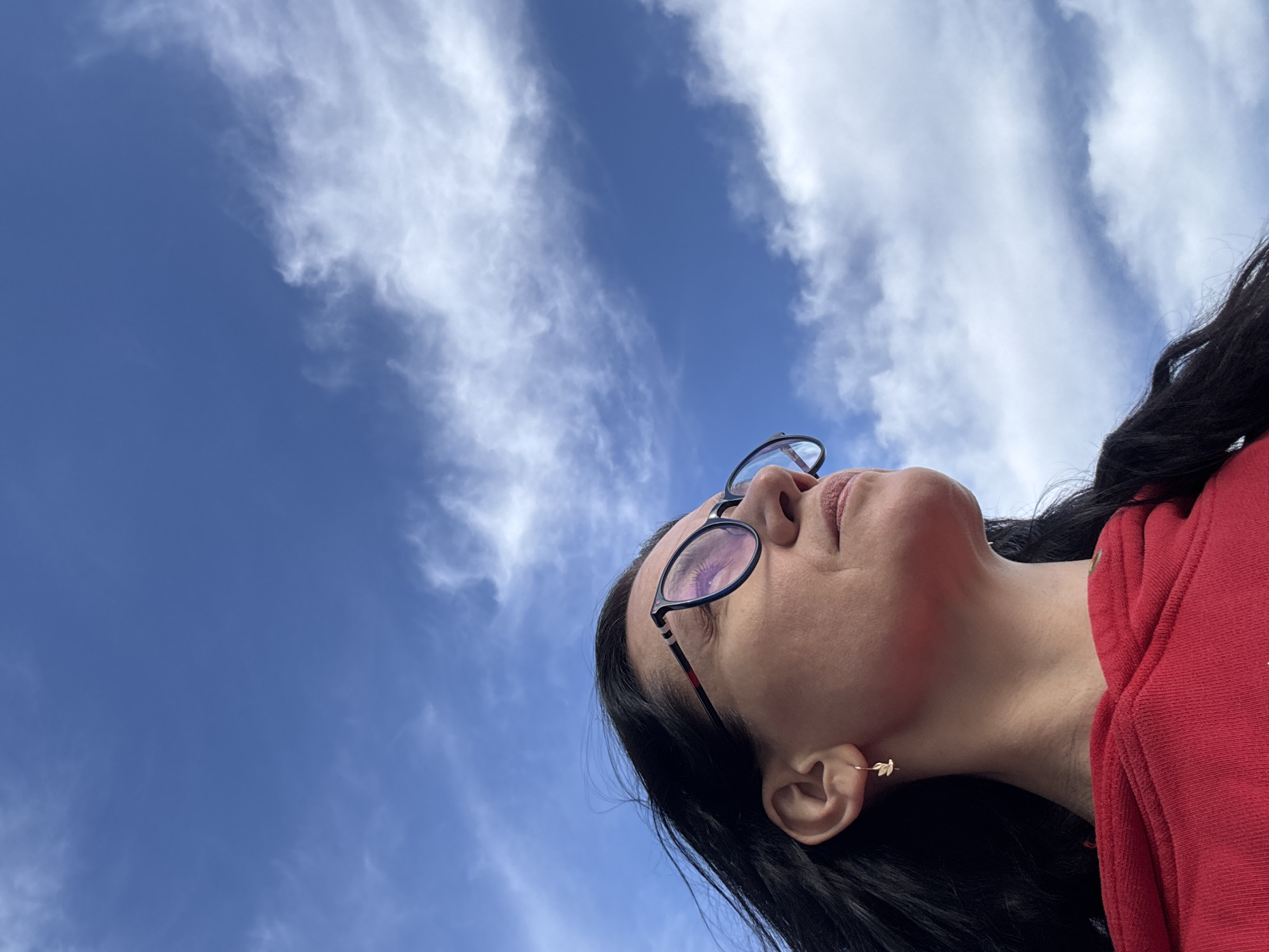 A low-angle shot of a woman with dark hair and glasses looking up at a blue sky with scattered white clouds. She is wearing a red hoodie and has a gold leaf earring. The location is unidentifiable as Chaikovsky, Russia based on the visible details. The focus is on the woman's face and the sky above her.