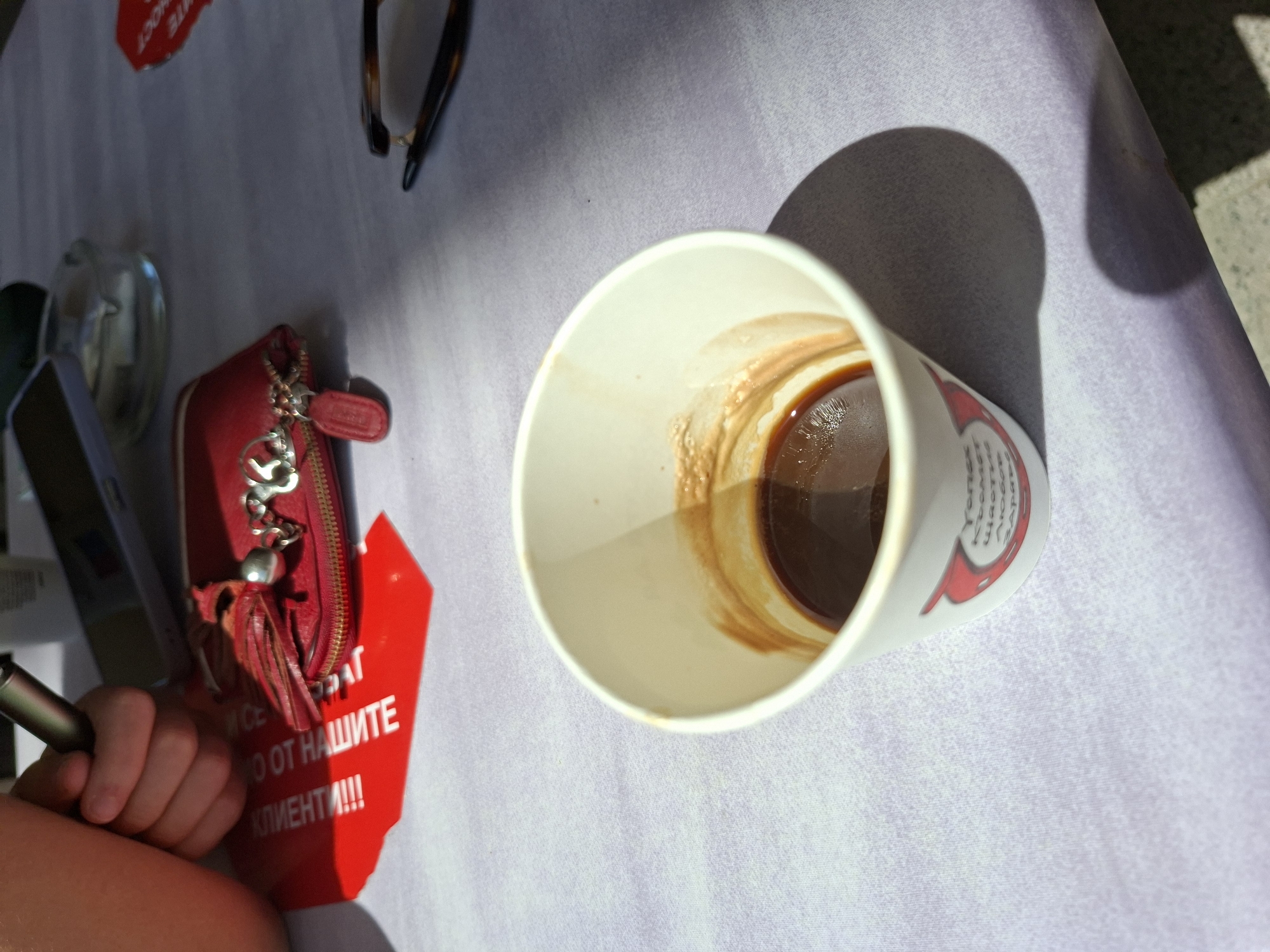This high-angle image depicts a casual outdoor scene, likely at a cafe or restaurant in Plovdiv, Bulgaria, bathed in bright sunlight as indicated by the sharp, distinct shadows cast by objects. The setting includes a table covered with a light-colored, textured tablecloth, possibly light grey or lavender.
The primary subject is a white paper cup, partially empty, containing a dark liquid, likely coffee, with noticeable residue on its inner walls. To the left of the cup, a red zippered wallet or small purse with a silver charm and a red tassel is visible. A light-skinned human hand, holding a silver pen or stylus, is seen next to the wallet. Further to the left, parts of a smartphone and a reflective ashtray are present. A pair of tortoise-shell sunglasses rests on the table towards the upper right. The far background suggests a darker, possibly paved outdoor surface.
The arrangement of items like a coffee cup, wallet, phone, and sunglasses implies a person is taking a relaxed break or enjoying a casual outing.
Visible text on a red card partially obscured beneath the wallet reads "ОТ НАШИТЕ КЛИЕНТИ!!!" (from our clients!!!), with preceding words largely unreadable. The paper cup also bears text in Cyrillic script, with fragments such as "ТОПЛ" (likely 'warm'), "ЛЮБОВ" (love), and "ЗАРА" (likely 'charge' or 'energy') visible. The Cyrillic script confirms the Bulgarian language context provided.