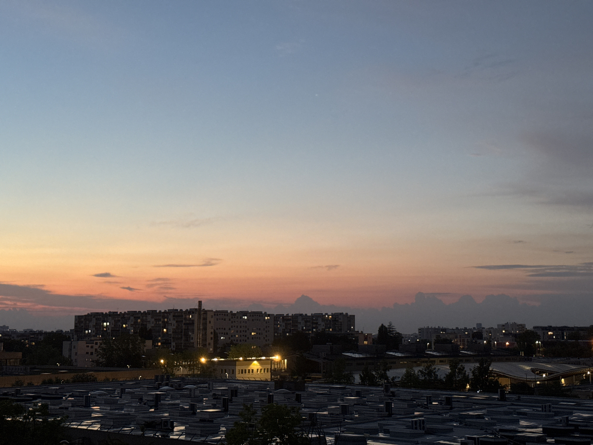 A dusky sky transitions from a light blue at the top to orange and pink hues near the horizon. Silhouetted buildings, including apartment blocks with illuminated windows, are visible in the mid-ground. In the foreground, a dense array of flat-roofed structures with visible ventilation units and pipes occupies the scene. The environment appears to be an urban industrial or residential area during twilight.