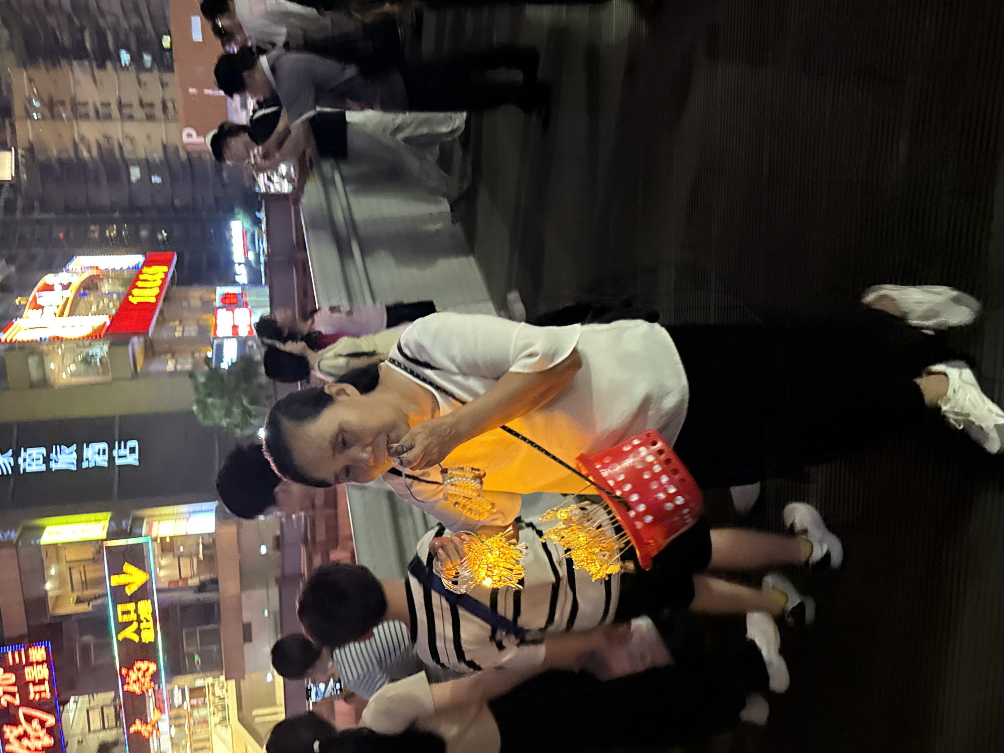 A woman is walking at night in Yuzhong District, China, holding a red basket filled with small, glowing items, possibly lanterns or sparklers. She is wearing a white shirt and black pants, and appears to be in her late 50s or early 60s. Several other people are in the background, blurred due to motion. The environment appears to be a street or pedestrian walkway, with illuminated signs and buildings visible in the background. The overall scene suggests a festive or commercial atmosphere.