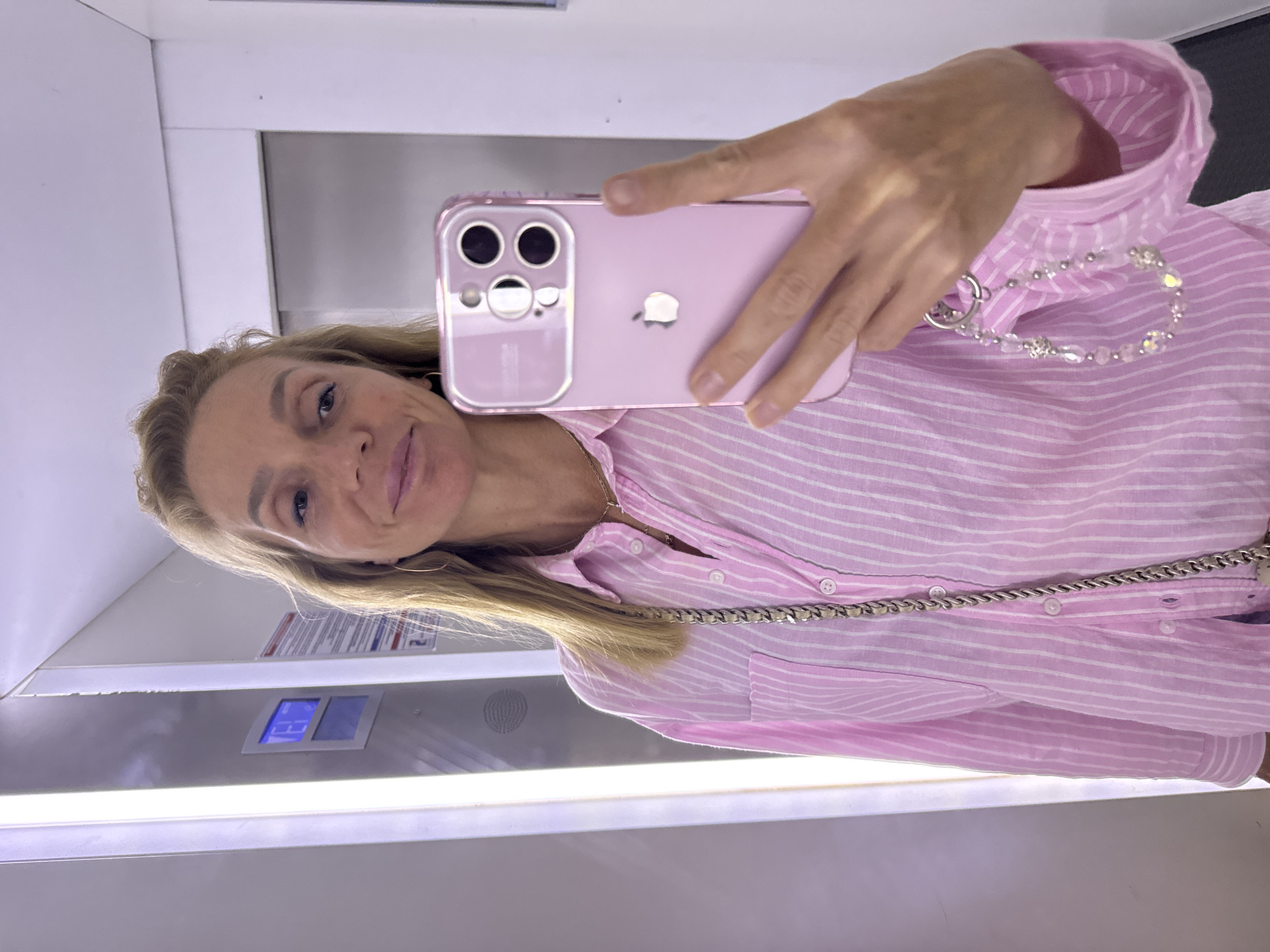 A woman takes a selfie in an elevator. She has blonde hair and is wearing a pink and white striped shirt, a necklace, and hoop earrings. She is holding a pink iPhone with a clear case and a beaded charm. The elevator is dimly lit with fluorescent lights on the left wall and a closed door on the right. There is a blue digital display on the wall, and some signage. The overall scene is mundane and typical of an urban environment.