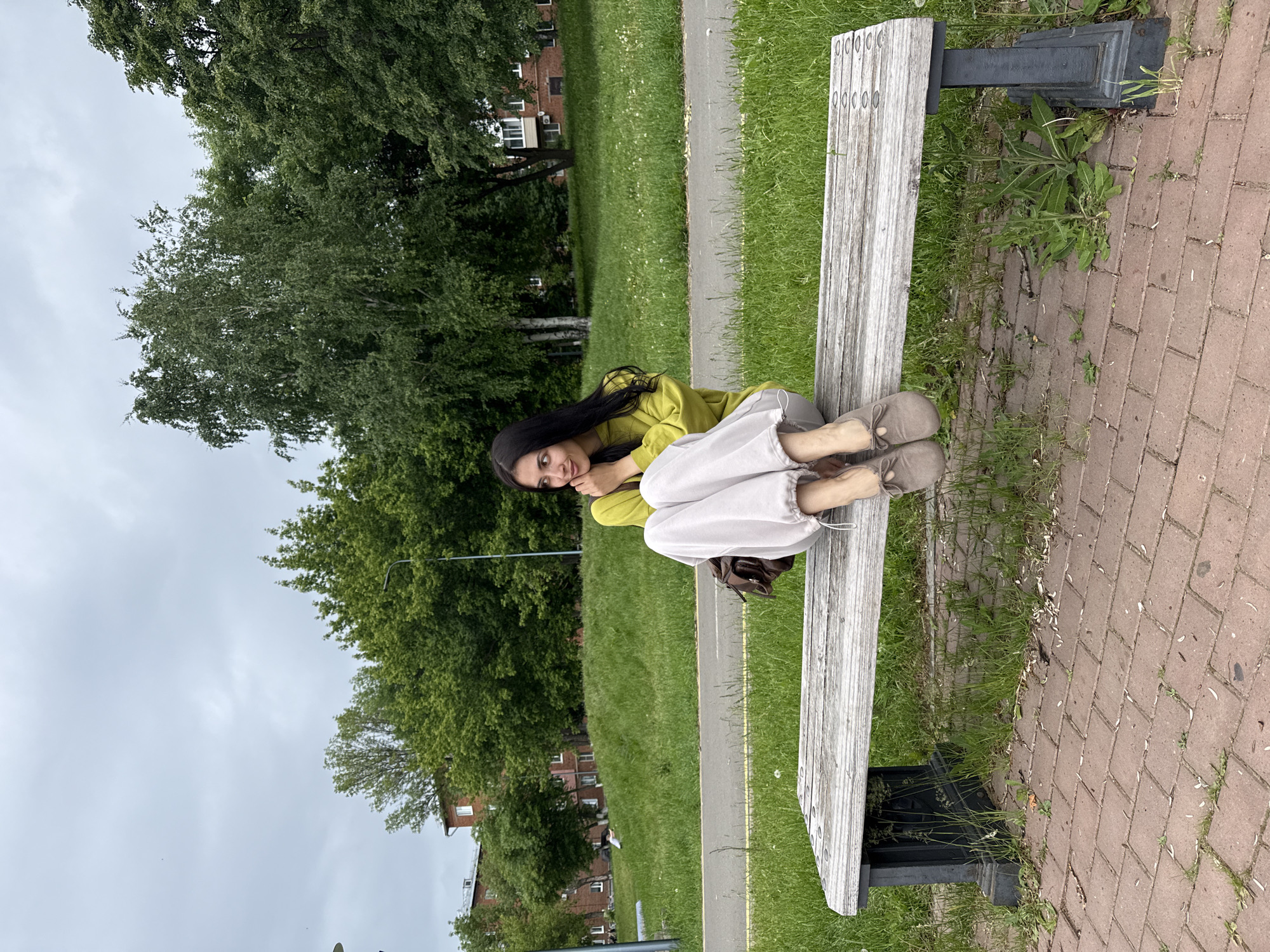 A young woman is captured sitting casually on a weathered wooden park bench in an outdoor green space. She has long, dark hair and is dressed in a chartreuse (lime green) long-sleeved top and light-colored, loose-fitting pants, complemented by light brown slip-on shoes. She is looking towards the viewer with a soft, pleasant smile, her body slightly angled, and her knees bent in a relaxed pose.
The bench is made of light-toned wooden planks with dark metal supports, positioned at the edge of a reddish-brown brick-paved area. Adjacent to the bricks is a vibrant green grassy lawn, through which a narrow paved path runs. The background features abundant green foliage from numerous trees, suggesting a park or a heavily treed area. In the distance, partially obscured by the trees, are sections of multi-story buildings constructed with red bricks, indicating an urban or suburban environment. The sky above is overcast and light grey, typical of a cloudy day. The scene conveys a tranquil moment in a public park, located in Votkinsk, Russia. There is no visible text in the image.