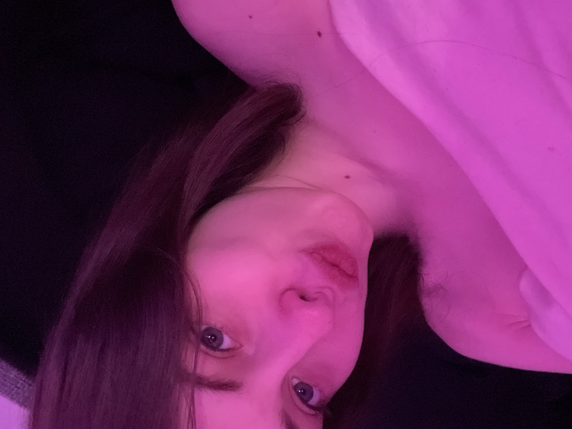 A young woman is visible in a close-up, angled shot. Her face is illuminated with a pink light, and she has blue eyes. She is wearing a light-colored garment, possibly a shirt. Her hair is brown. The background is dark. The location and additional details are not ascertainable from the image.