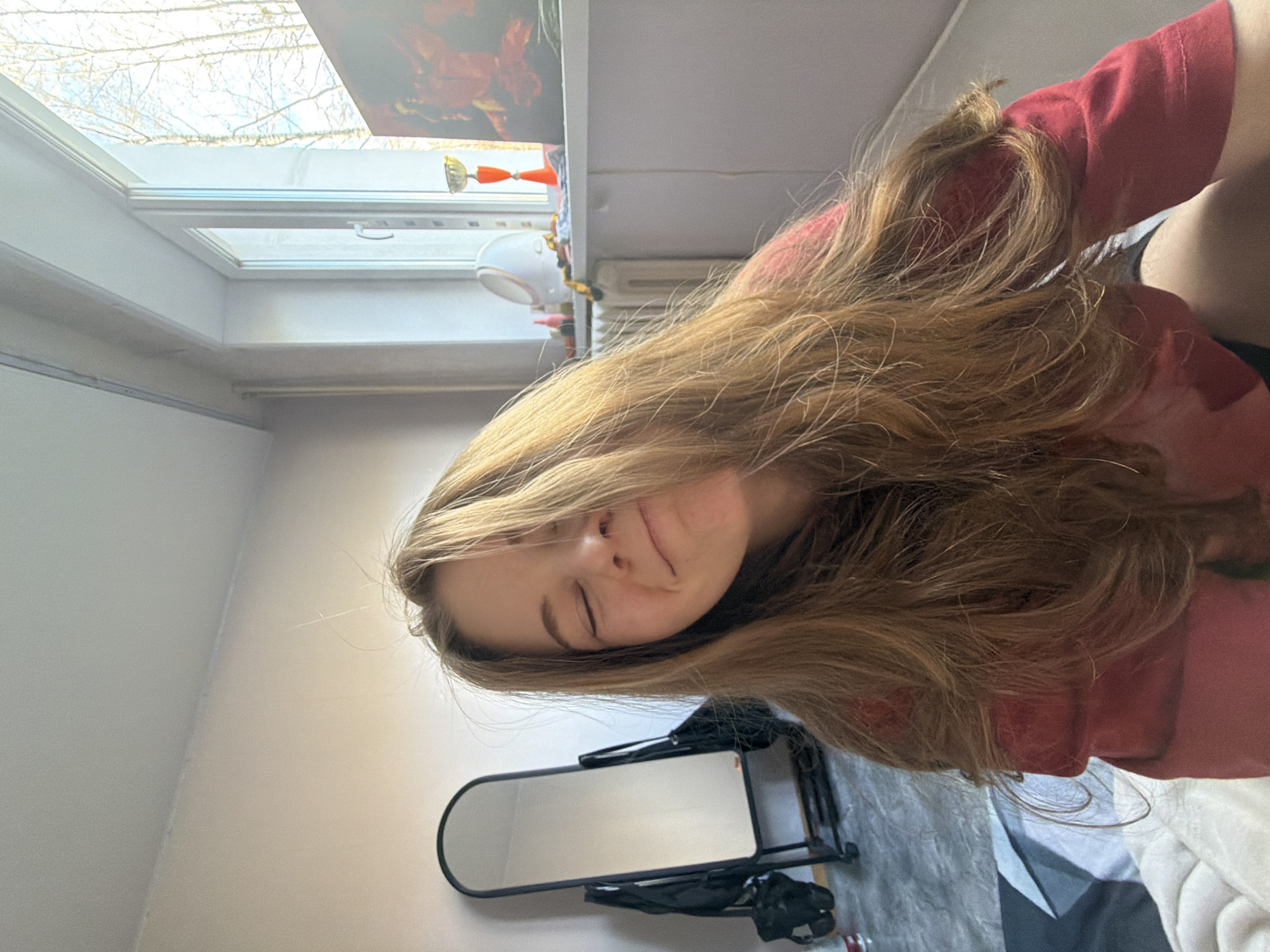 A young woman with long brown hair is lying down indoors, possibly in Perm, Russia, as suggested by the user prompt. She is wearing a red top. A mirror is visible on the floor. A window with leafless trees visible outside is in the background. Other items are seen near the window. The scene appears to be a bedroom or living space. The angle of the photo is taken from above.