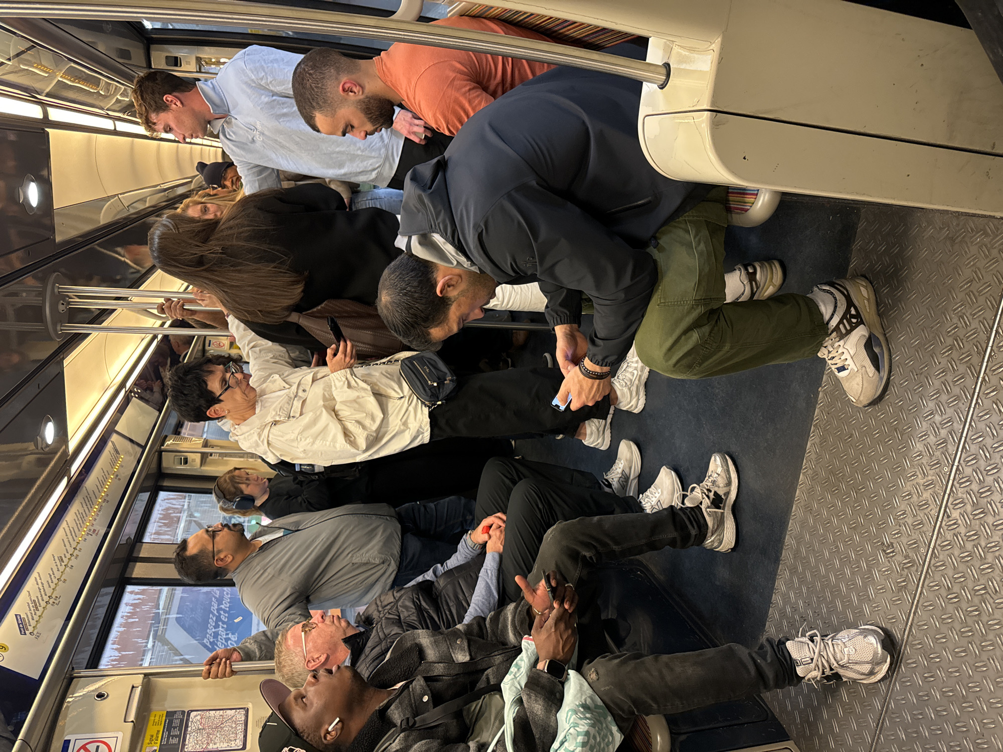 The image depicts the interior of a subway car, likely in Paris, France, based on the signage and design elements. Numerous passengers are visible, occupying both seating and standing positions. They exhibit varied postures and activities, with many using mobile phones, looking downward, or leaning against support bars. Clothing styles are diverse, ranging from casual to more formal attire. The interior of the subway car includes typical features such as handrails, informational signage (including a subway map), and patterned flooring. The lighting appears to be artificial, with overhead fixtures illuminating the scene. The overall atmosphere suggests a crowded and functional urban transportation setting.