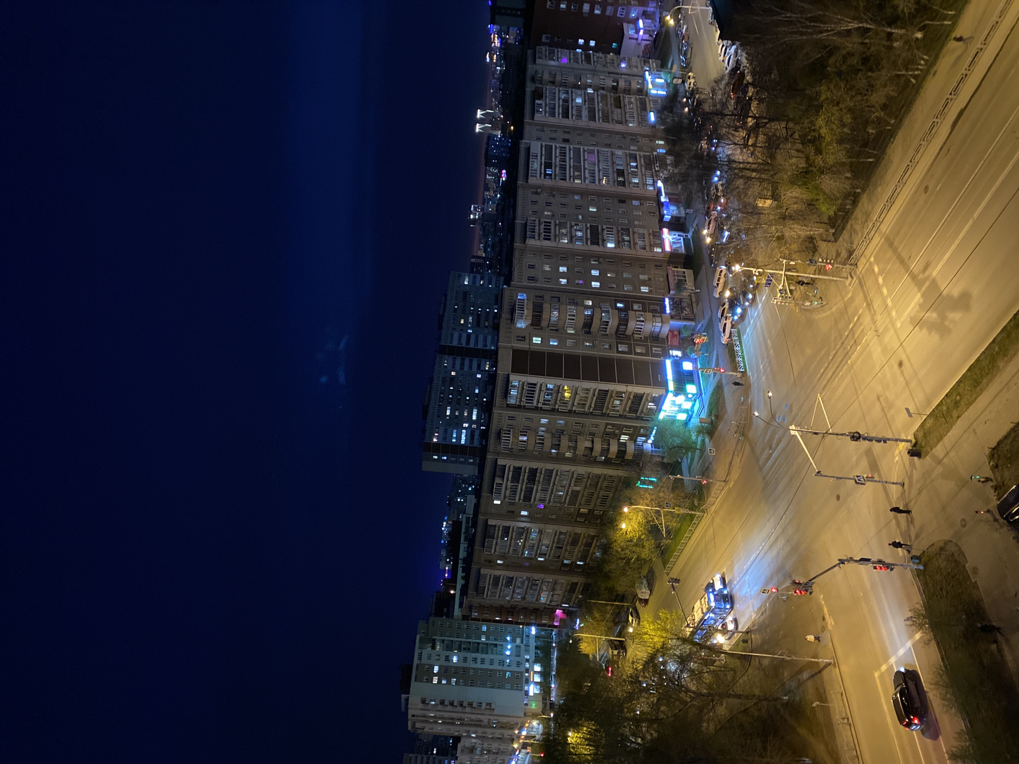 The image captures a high-angle, nighttime view of a city street and buildings in Yekaterinburg, Russia. The perspective looks down onto the scene, with a deep blue, almost black, night sky filling the upper left portion of the frame. In the distance, faint city lights twinkle on the horizon.
Along the right side and stretching into the middle ground, multi-story residential and commercial buildings line a broad street. Many windows in these buildings are lit, casting warm glows and suggesting ongoing activity within. Some buildings feature balconies, and various commercial signs, though unreadable, are visible on lower floors, glowing in shades of blue, green, and pink.
The foreground is dominated by a wide, multi-lane asphalt road that appears wet, reflecting the artificial light sources like a mirror. Several streetlights illuminate the road with a yellowish glow. A complex intersection is visible, equipped with multiple traffic light poles. Some traffic lights show red, while others are green or off. Several vehicles, including cars and what appears to be a bus or large van, are present on the road and at the intersection. Crosswalk markings are clearly visible on the pavement.
Scattered trees, mostly bare or with very sparse foliage, line the sidewalks and medians, suggesting it might be late autumn or early spring. A few pedestrians are visible on the sidewalks and crossing the street, appearing as dark silhouettes against the bright road. The overall atmosphere is that of a bustling city evening, illuminated by abundant artificial light sources against the dark night. No discernible text is clearly readable on any signs or objects within the image.