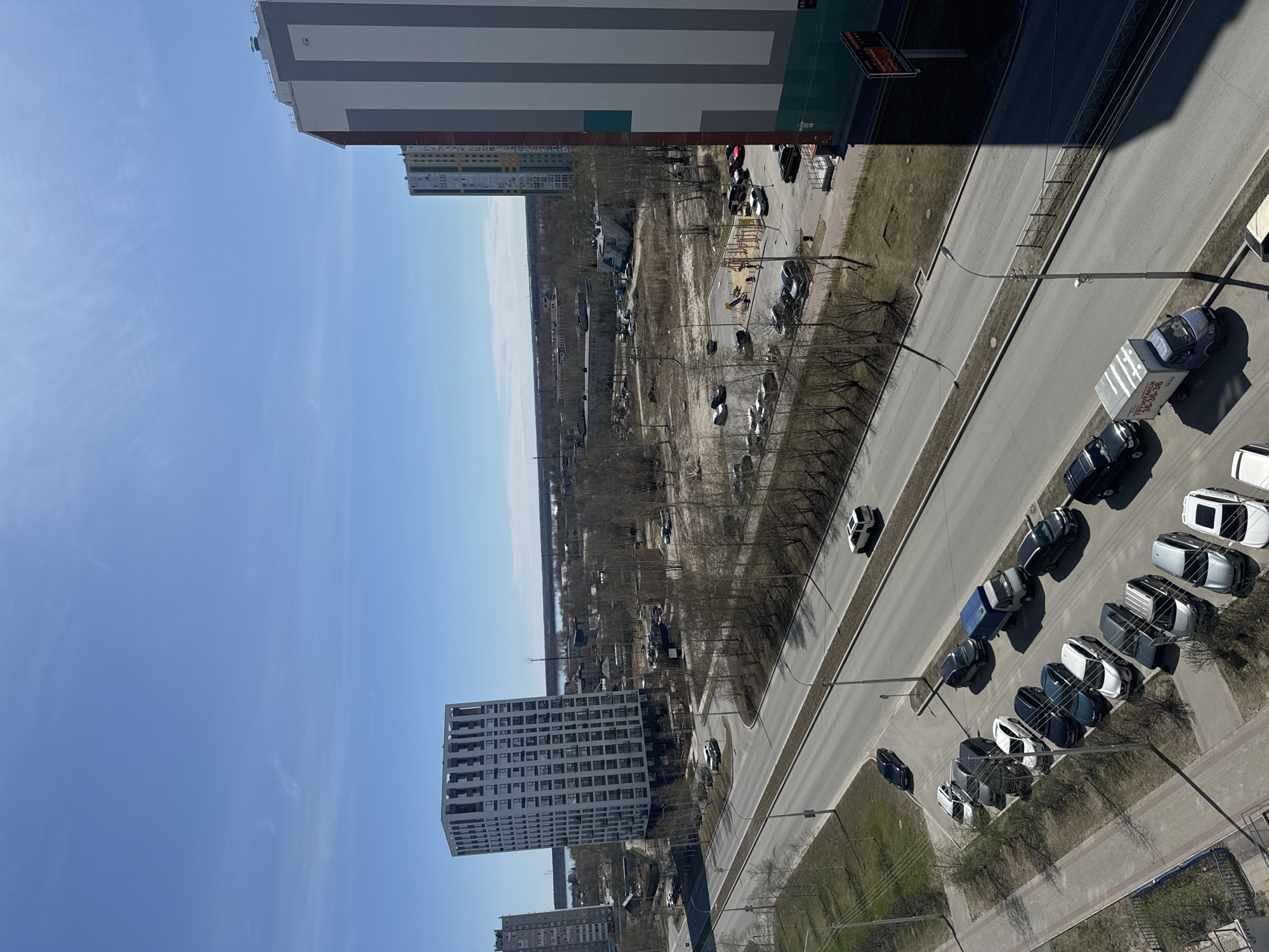 The image shows a high-angle view of a street and surrounding buildings in Surgut, Russia. Several apartment buildings are visible, ranging from mid-rise to high-rise structures with various color schemes and architectural styles. A road runs alongside a parking area filled with parked vehicles. Trees line the road and parking lot. In the distance, a more sparsely developed area with bare trees and what appears to be a body of water can be seen. The sky is clear and blue.