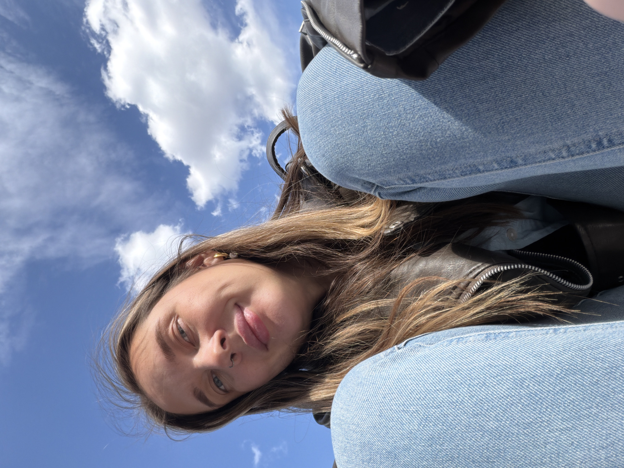 A woman is depicted from a low angle, her face and shoulders dominating the frame. She has fair skin, light brown hair with blonde highlights, and blue eyes. She is wearing a nose ring and a gold earring. Her expression is neutral, with a slight hint of a smile. She is wearing a dark leather jacket and blue jeans. Behind her, a bright blue sky is visible with scattered white clouds. This image was not taken in Moscow, Russia and there is no information about the location of the photo.