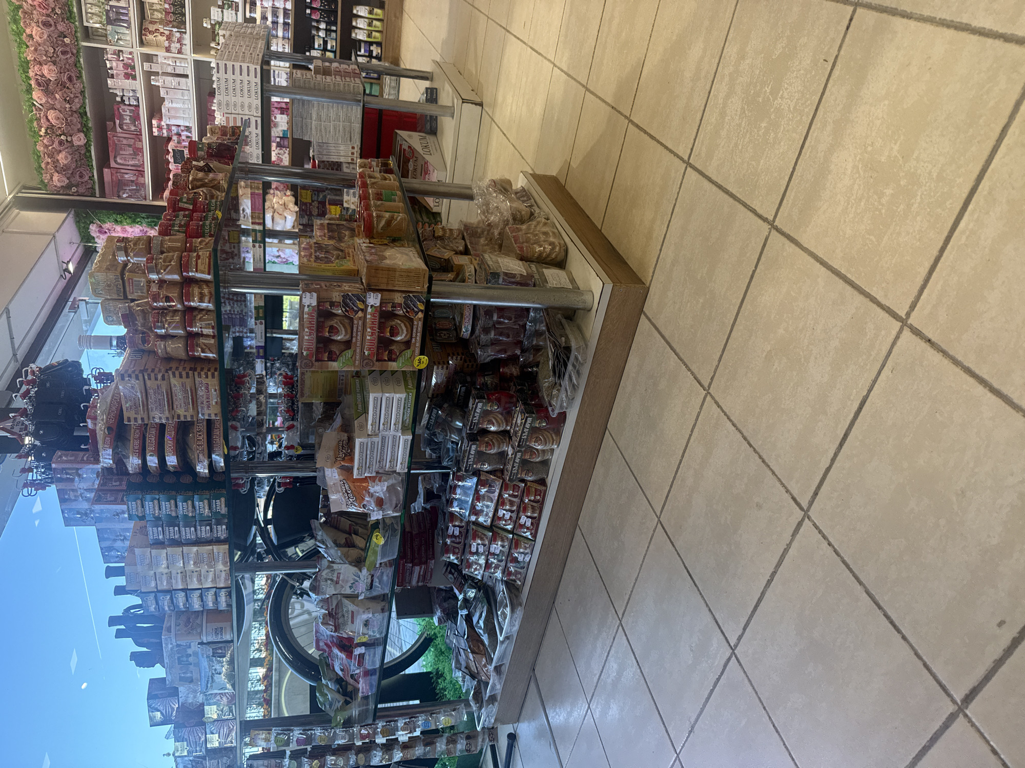 The image shows a retail display of various food items and products. The display is set up in a store with tiled flooring. On the shelves, there are boxes of spices, dried fruits, nuts, and other packaged goods. Some of the packaging is in Bulgarian. The store appears to be well-stocked, with products arranged in an organized manner. In the background, there is a large window offering a view of the sky and some greenery outside. The overall impression is that of a local market or specialty food store. This scene is consistent with a shop located in Varna, Bulgaria, given the Bulgarian text visible on some of the products.